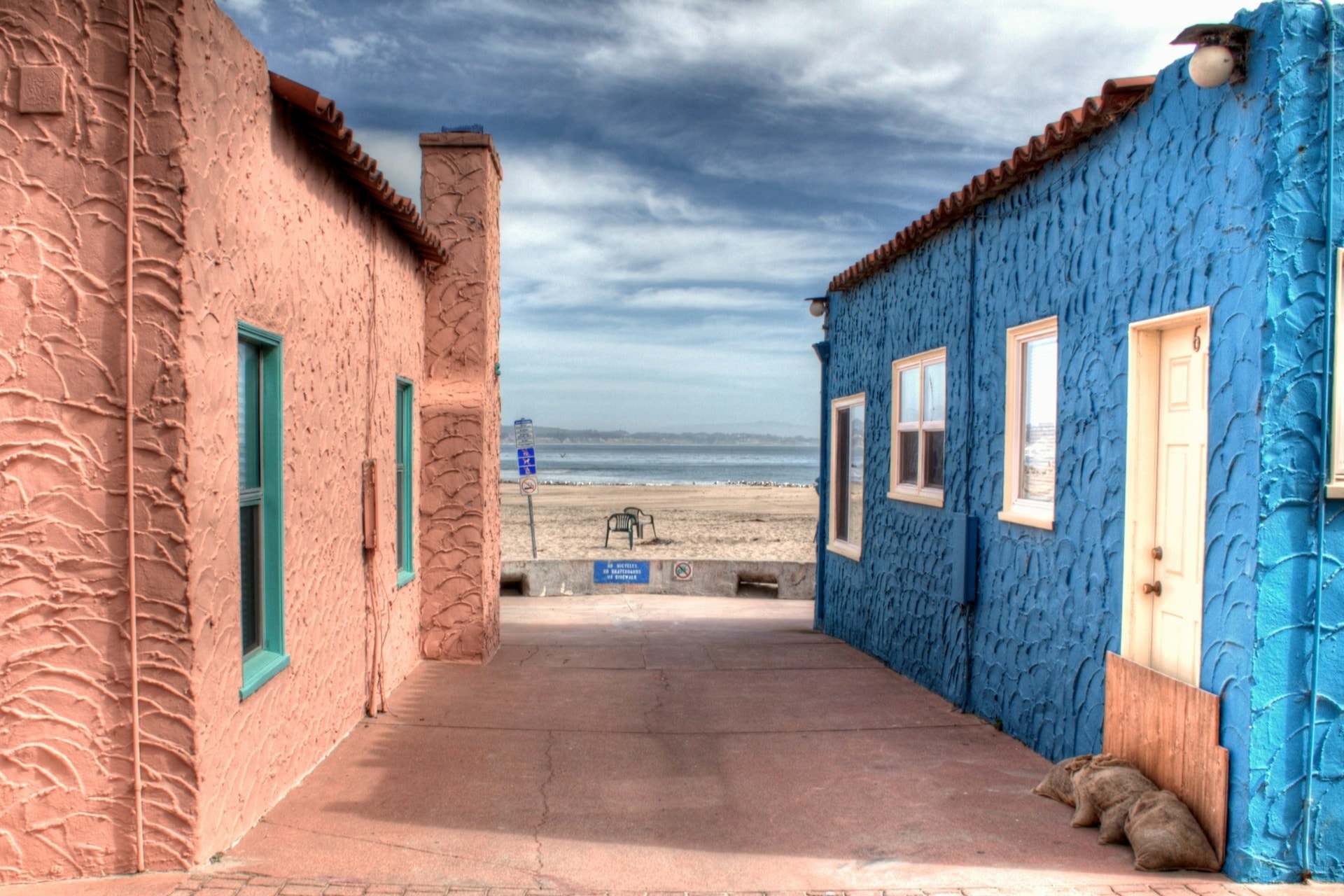 A narrow alleyway between an orange house and a blue house leading to a beach