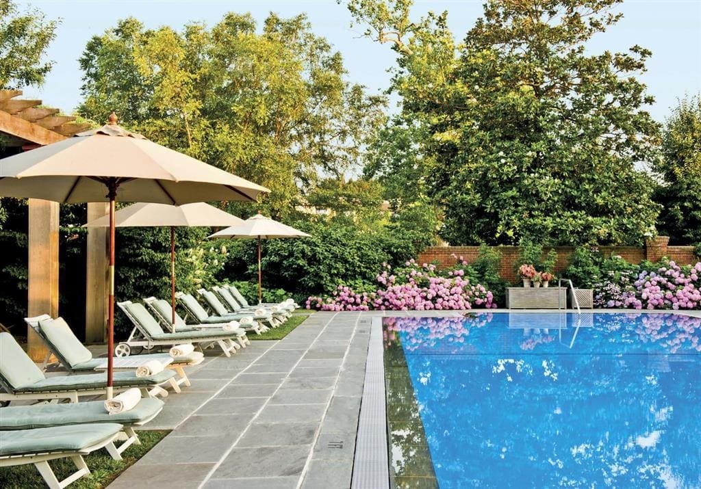 light-green loungers, green trees and hedges, and pink flowers surround a pool on a sunny day