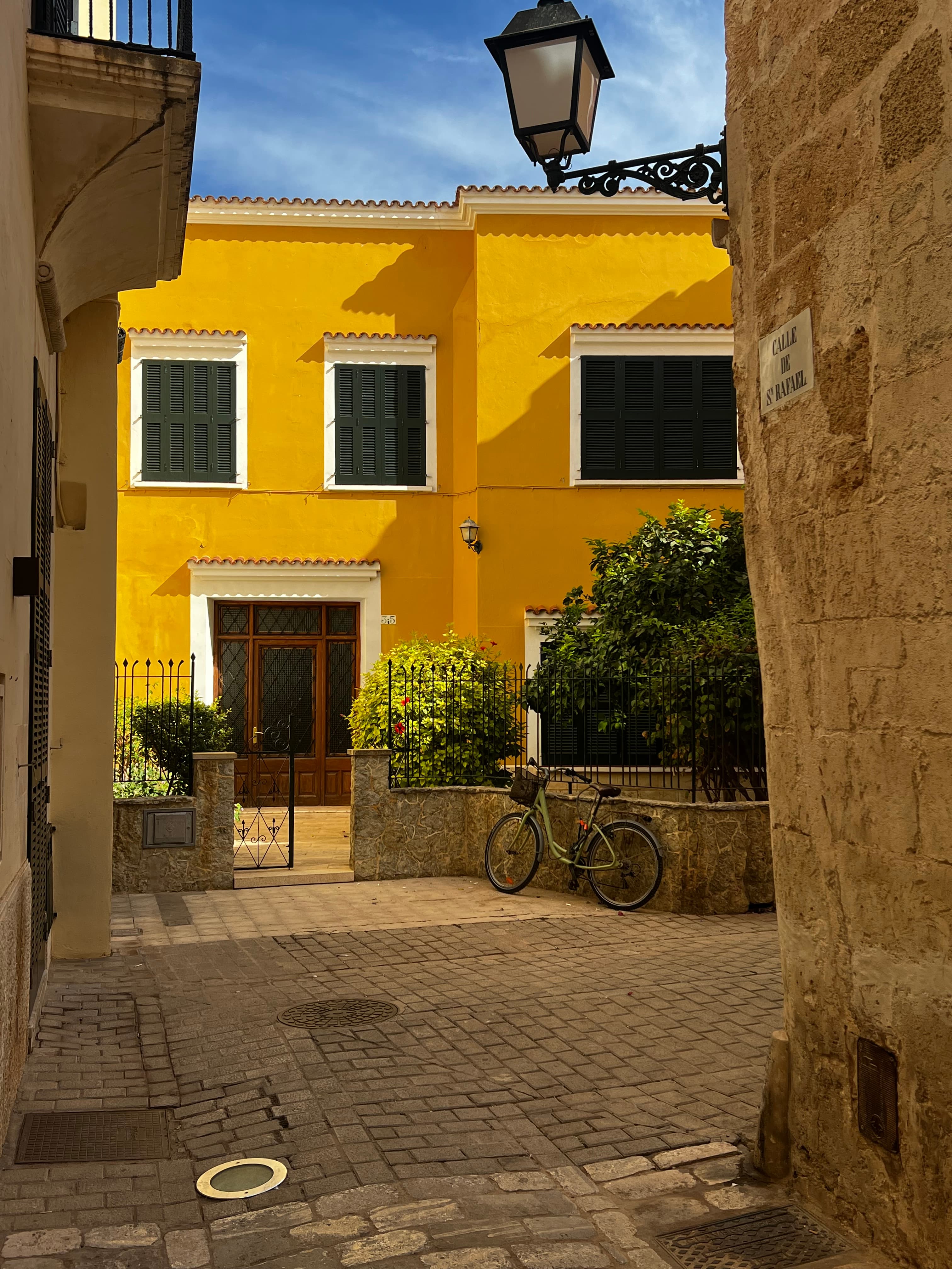 A beautiful picture of a yellow house surrounded by green bushes, stone walls, a bicycle and lanterns.