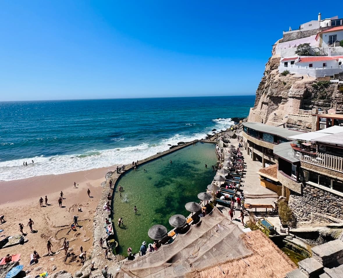 A resort with a green tinted swimming pool and beach with people walking around and the ocean in the background
