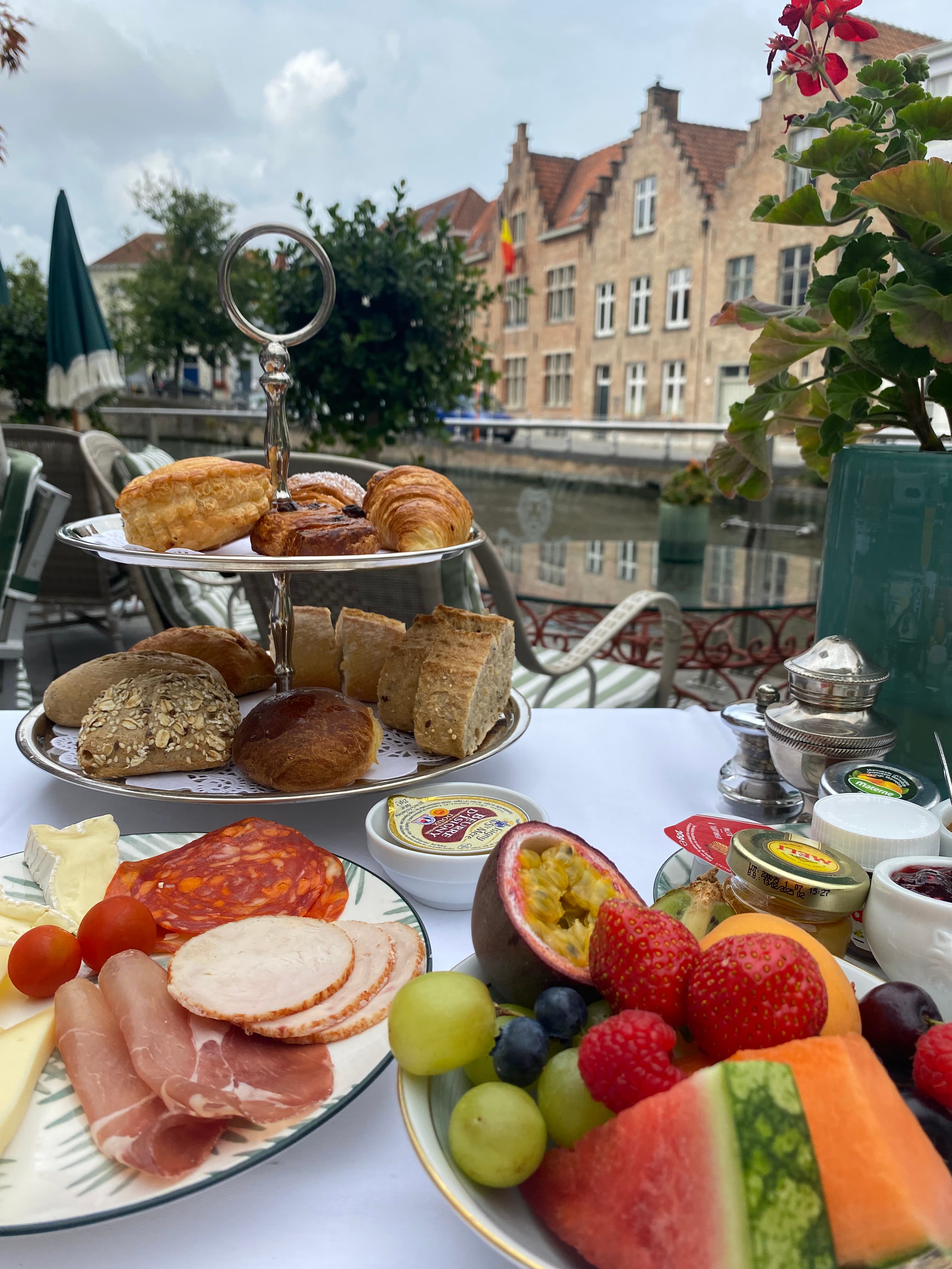 a table with a view of a canal and row houses with fruit, cheese, bread and meat plates on it