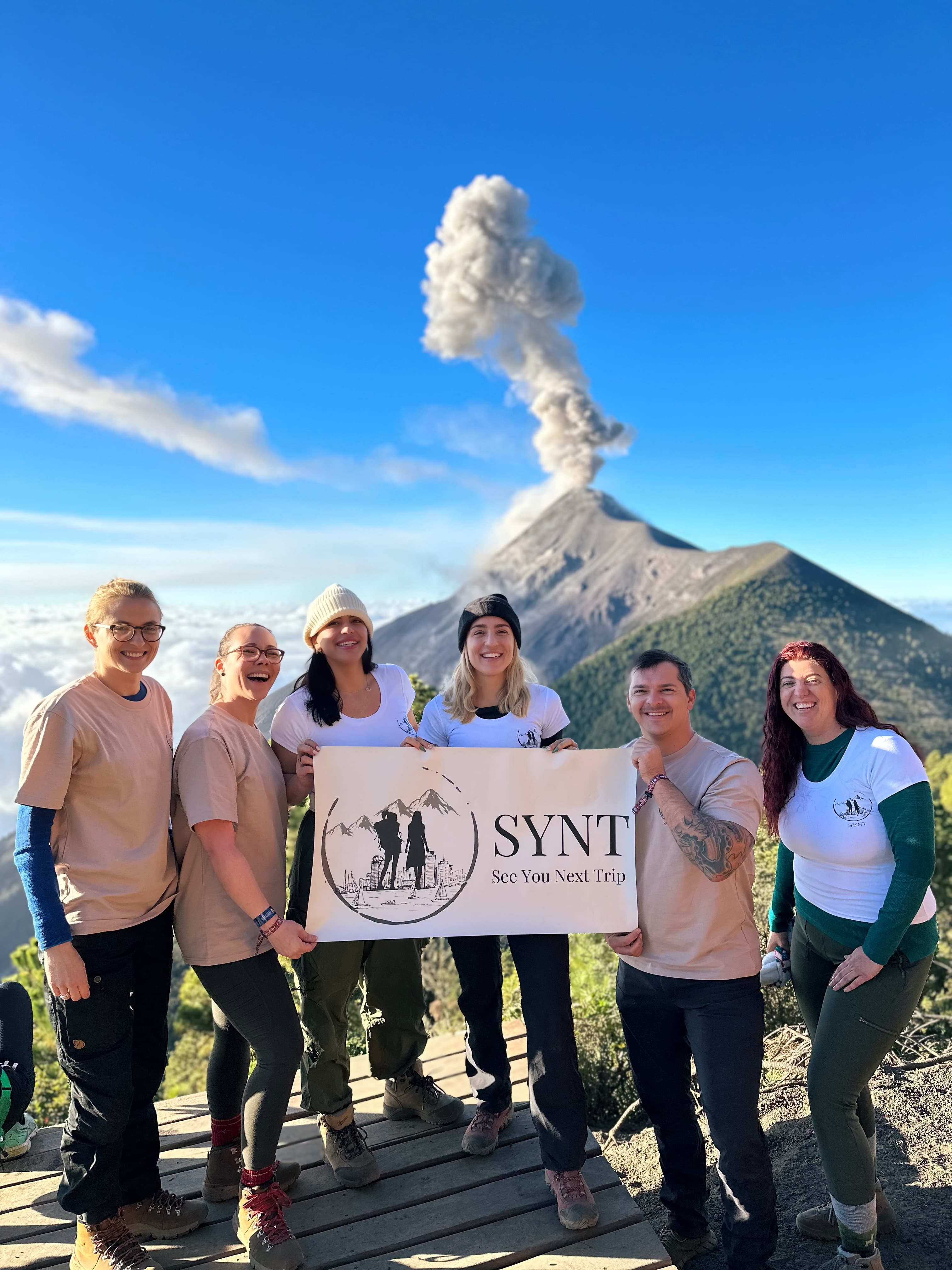 A group photo of six people holding up one sign and standing in front of a volcano