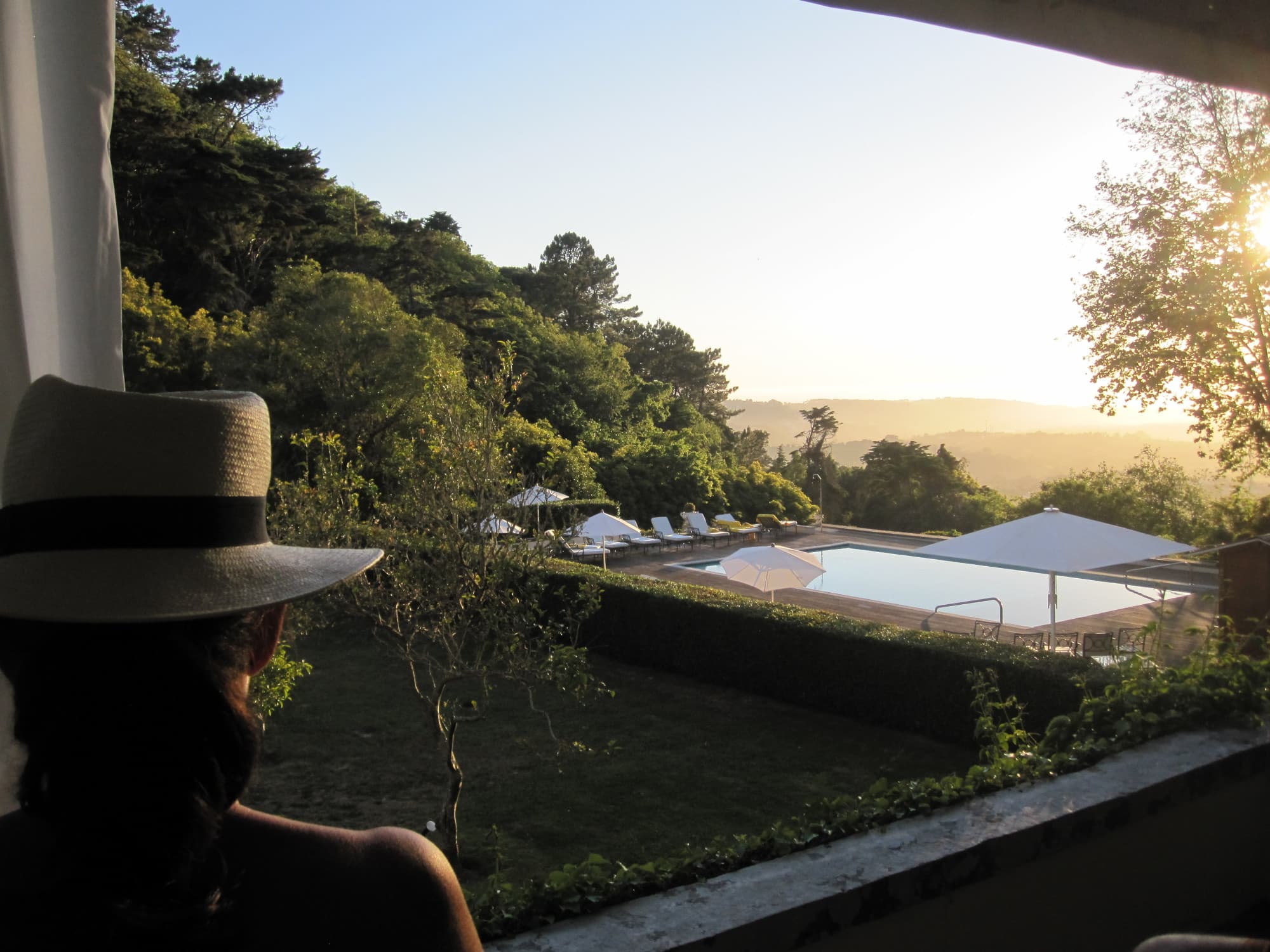A woman in a hat overlooks an infinity pool on a jungly landscape.