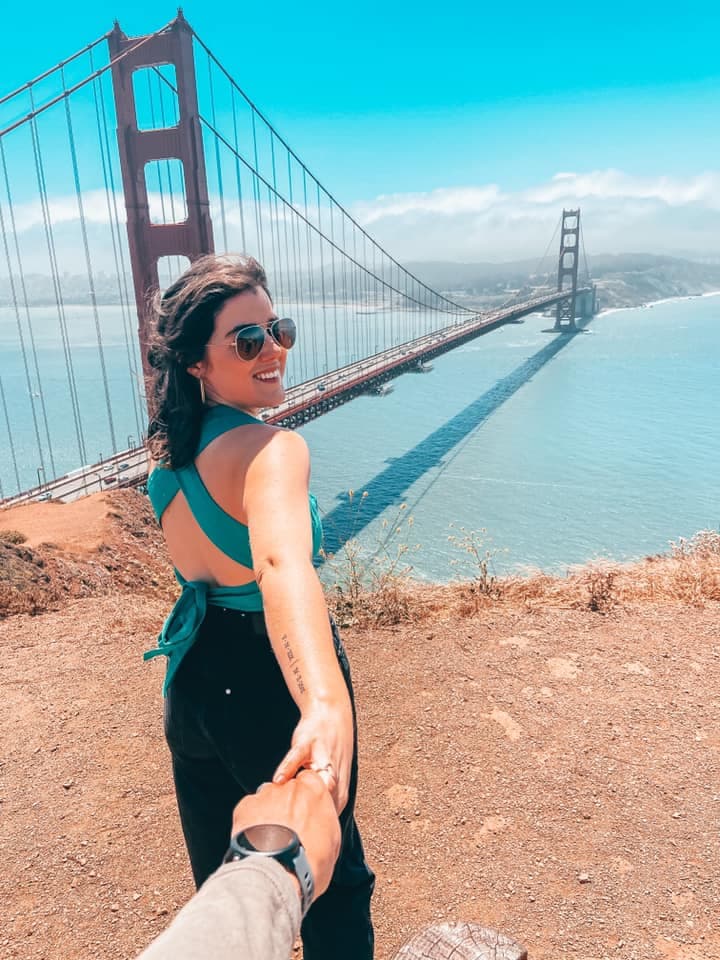 A girl standing in front of the Golden Gate Bridge in a blue top and black jeans, looking back at someone holding her hand