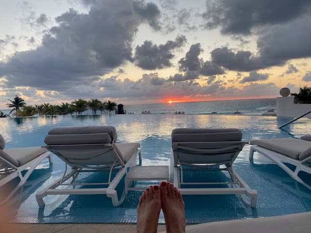 Feet positioned in front of two lawn chairs against a swimming pool that looks out to the sea and a sunset around scattered clouds and palm trees
