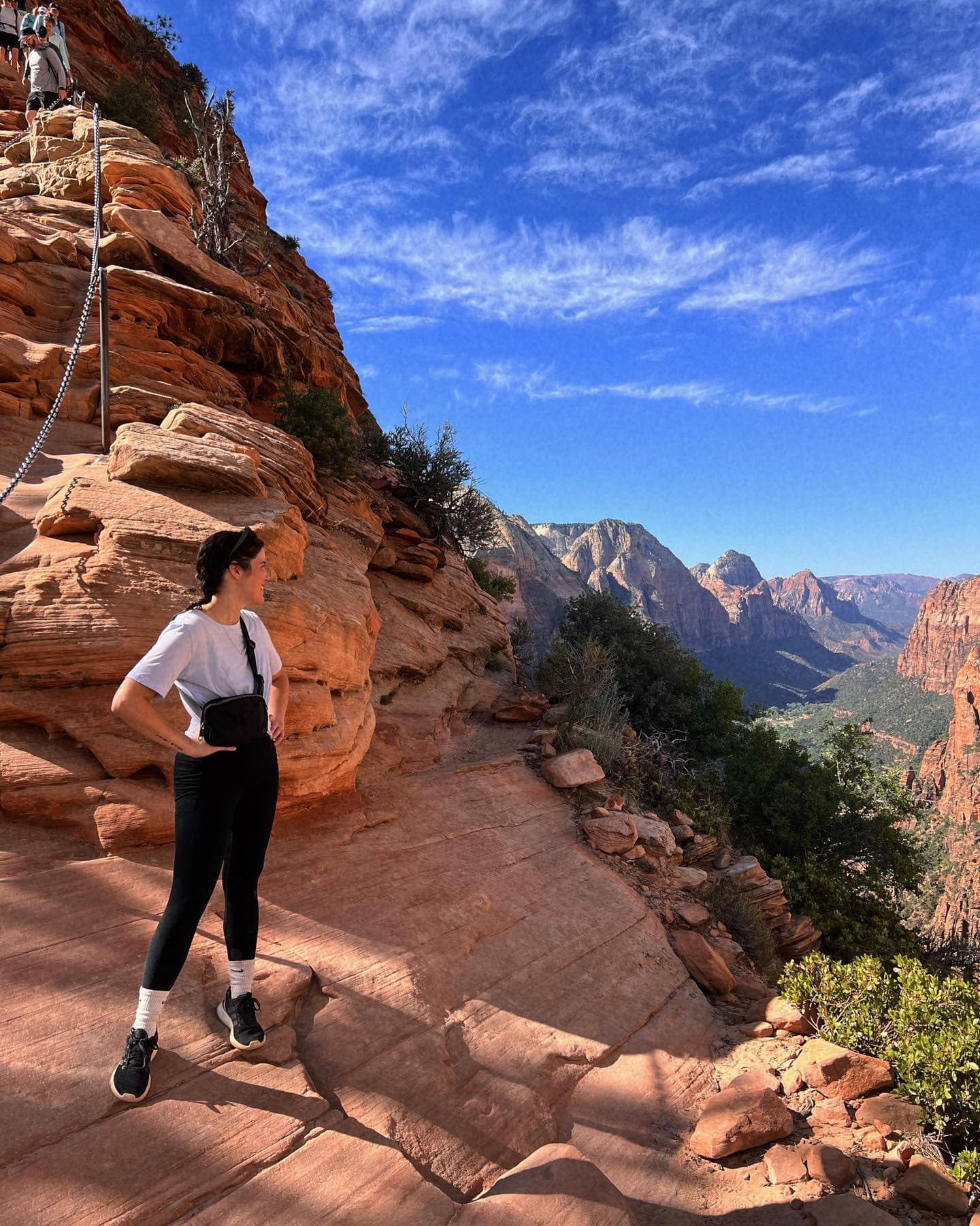 A girl standing on a clay-colored, rocky path in the middle of the mountains with a blue sky in the background
