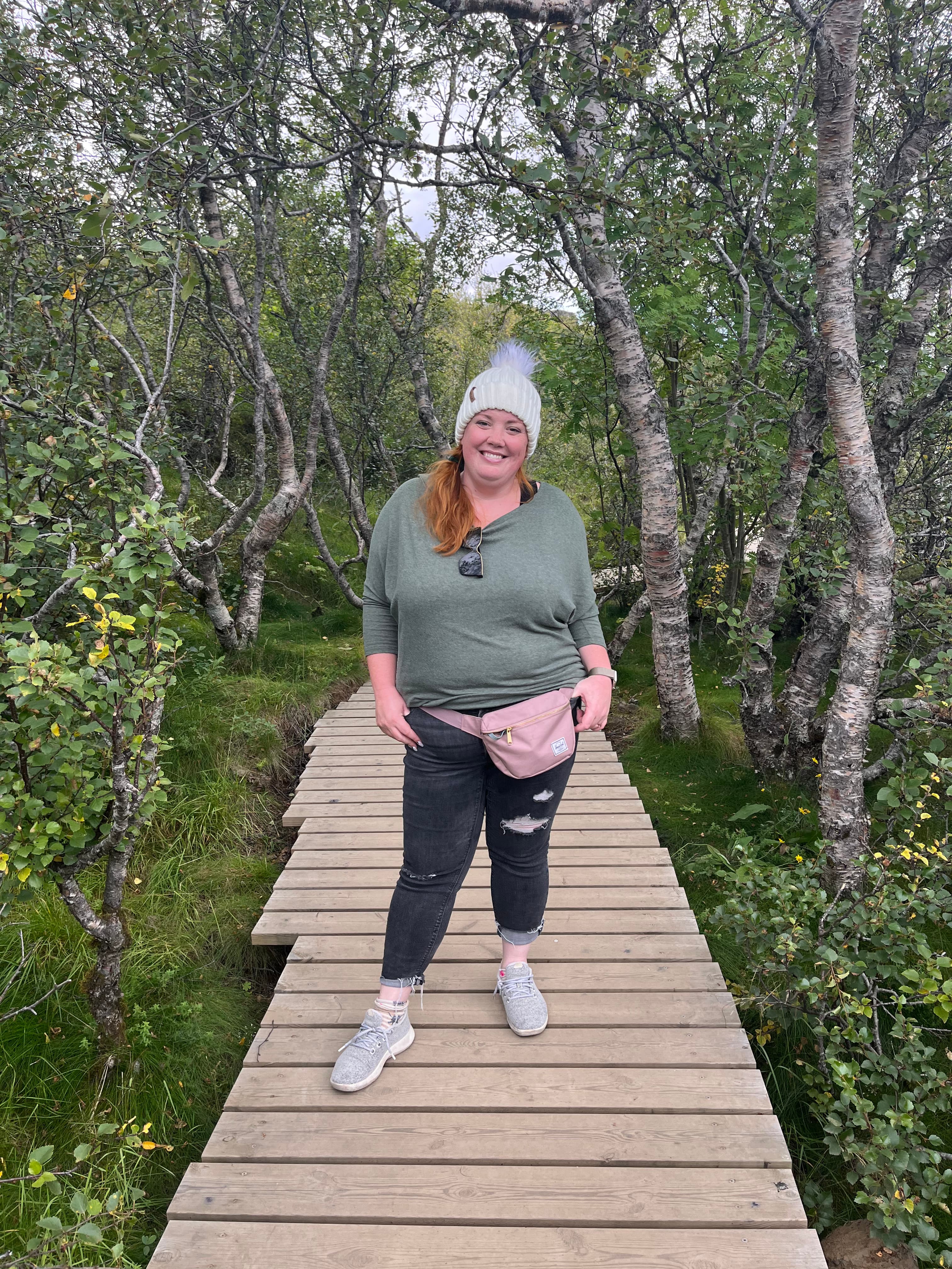 Emyli wearing a white beanie, green top, pink fanny pack and black pants on a wooden pathway surrounded by green trees and bushes