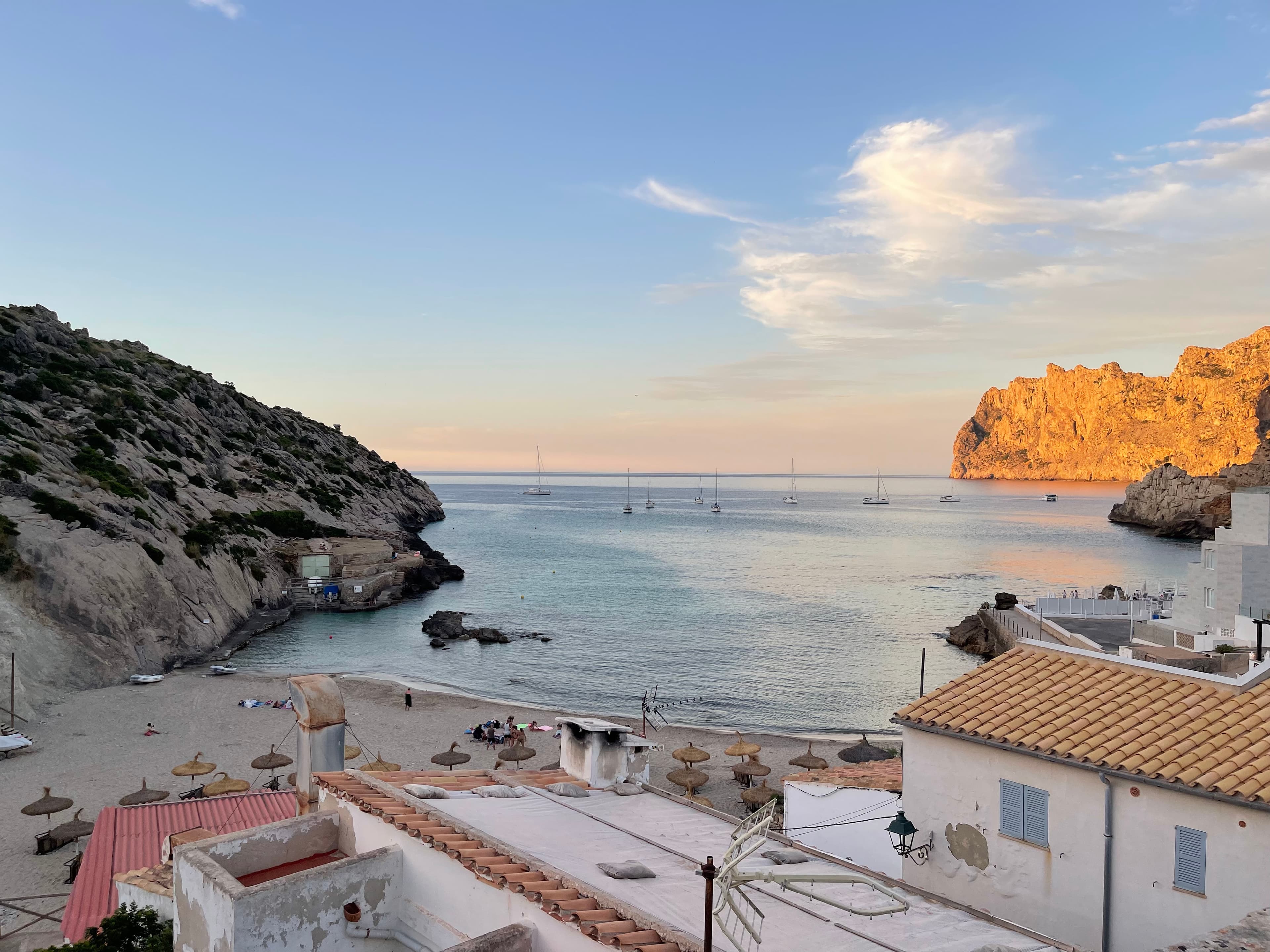 A view of the beach with terracotta-roofed and white buildings, blue sea and rocky cliffs in the background.