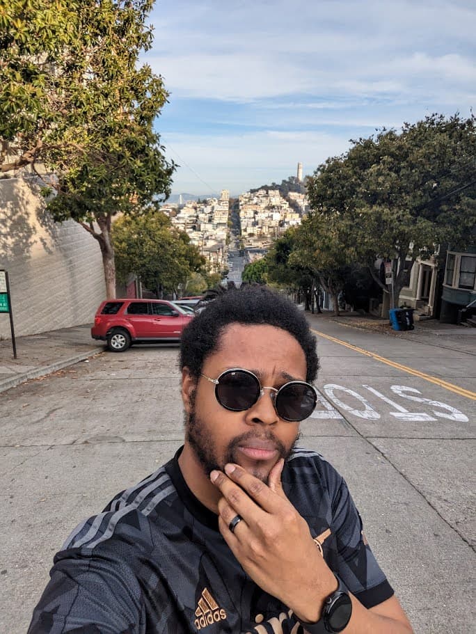 Norris posing for a selfie on a neighborhood street with a red car and city in the background