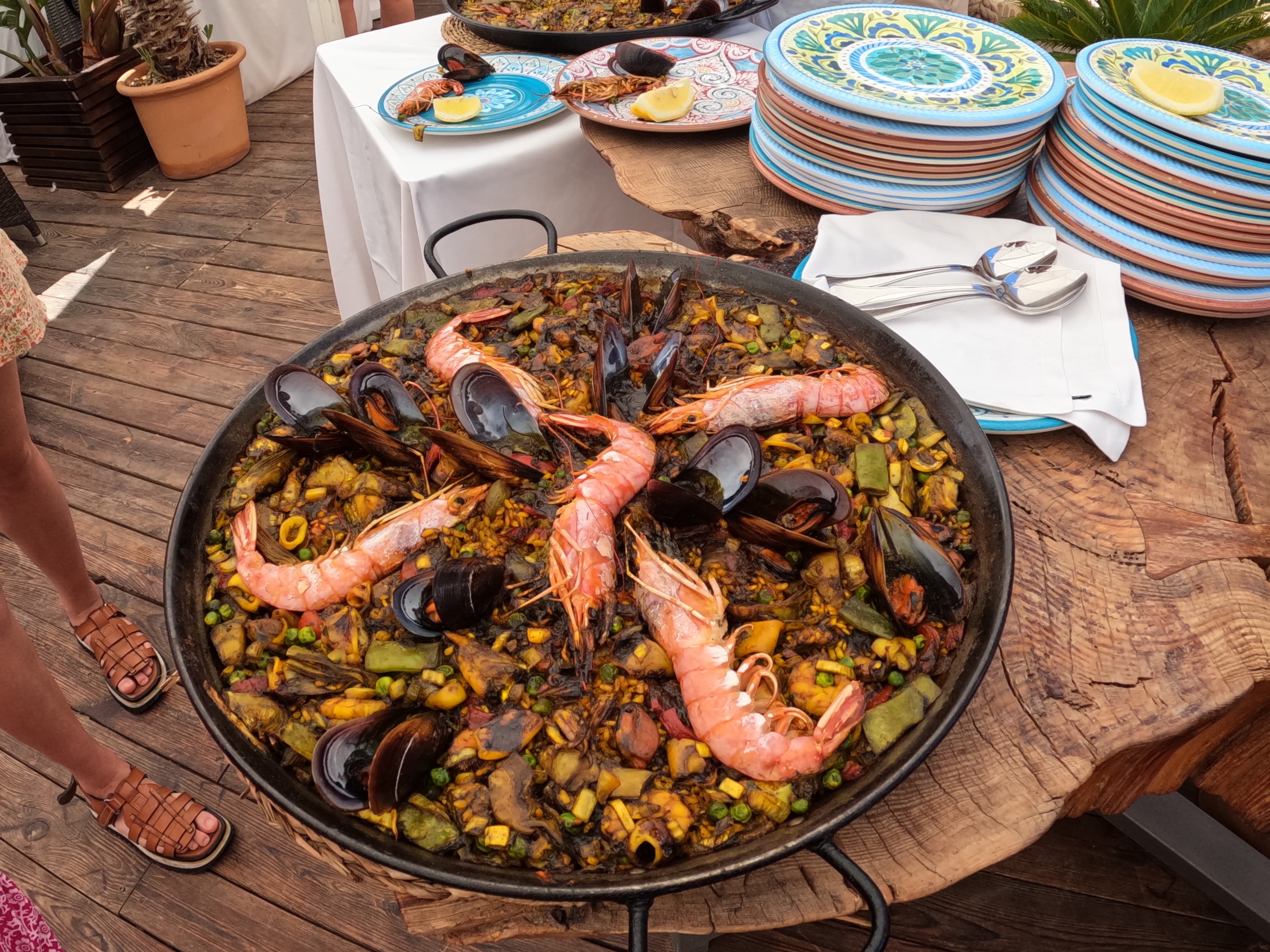Paella on a wooden table surrounded by colorful plates, spoons and tables in the background.