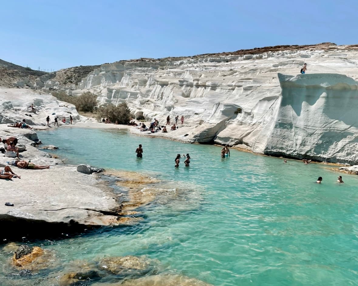 People swimming in a turquoise lake surrounded by white rocks and clear blue skies