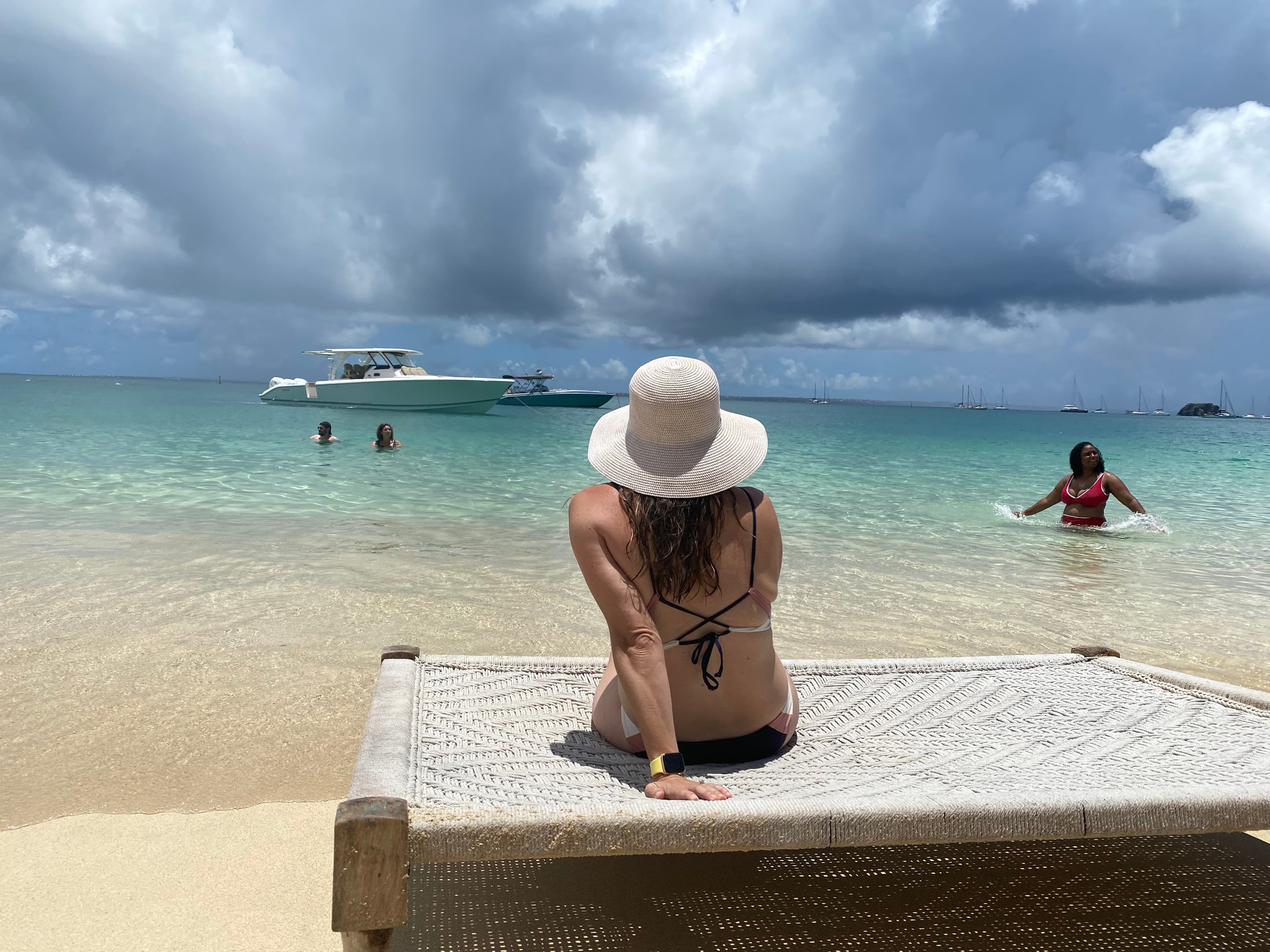 Latha wearing a black bikini and white hat looking out to the crystal blue water and boats from the beach on a cloudy day.