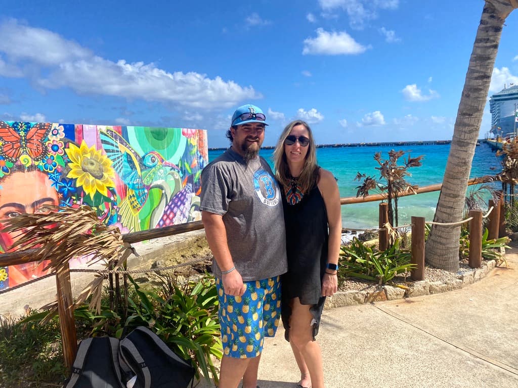 Picture of Amanda in a black dress with her husband in front of a colorful mural and sea view