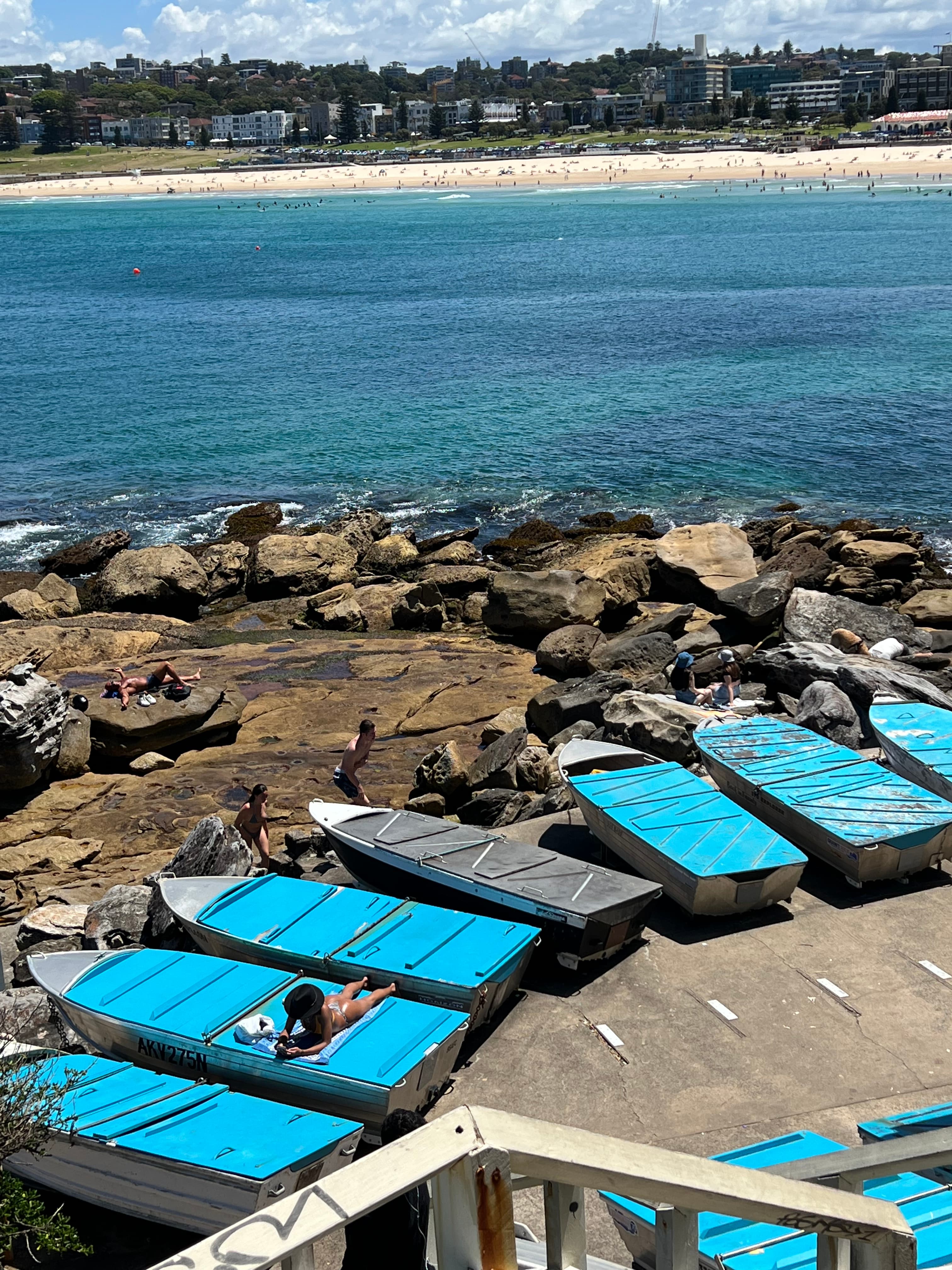 Blue boats lined up against a rocky shoreline with blue water and a white sand beach in the distance