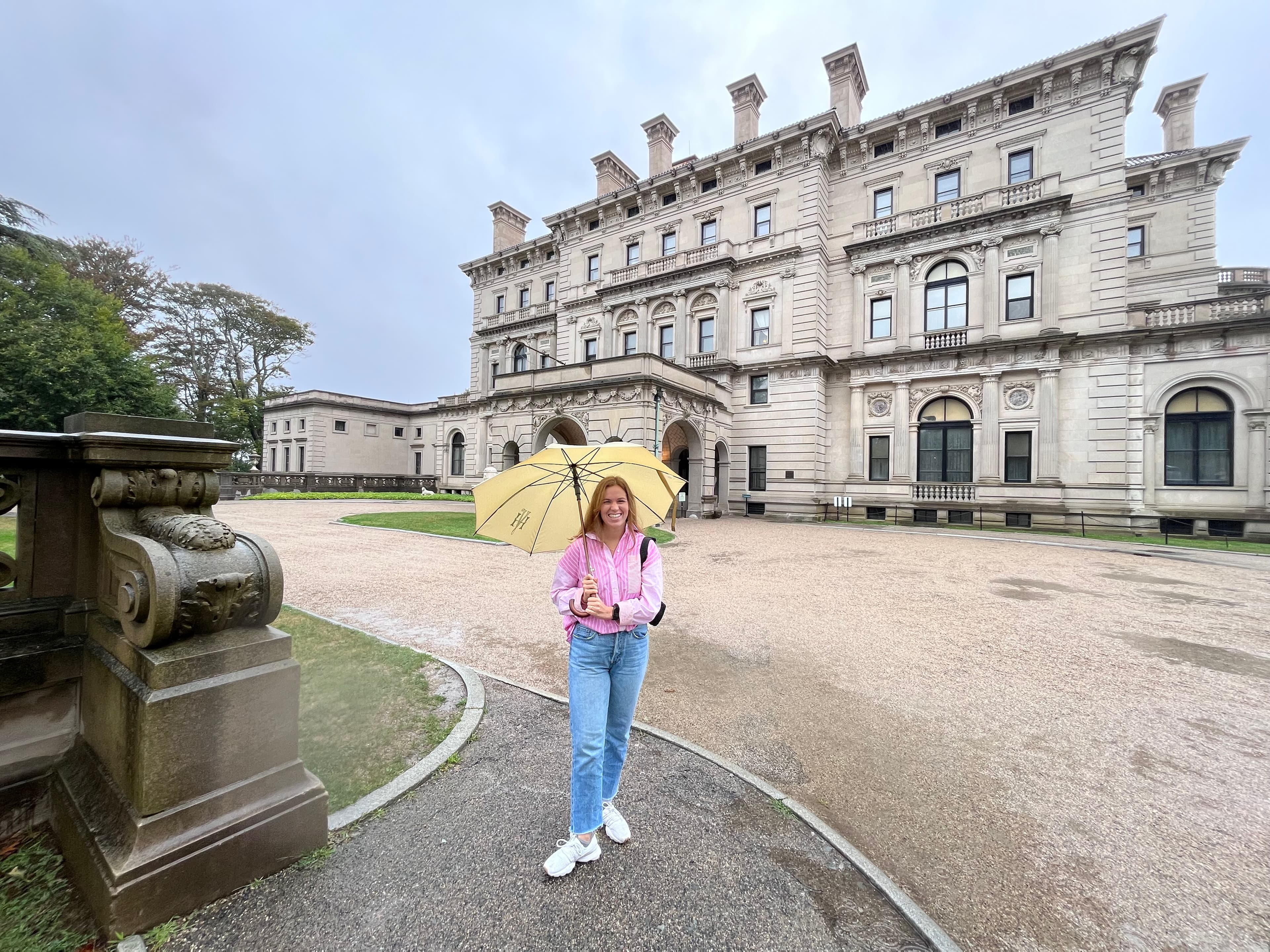 Sedona wearing a pink top and blue jeans holding a yellow umbrella in front of an old historic building