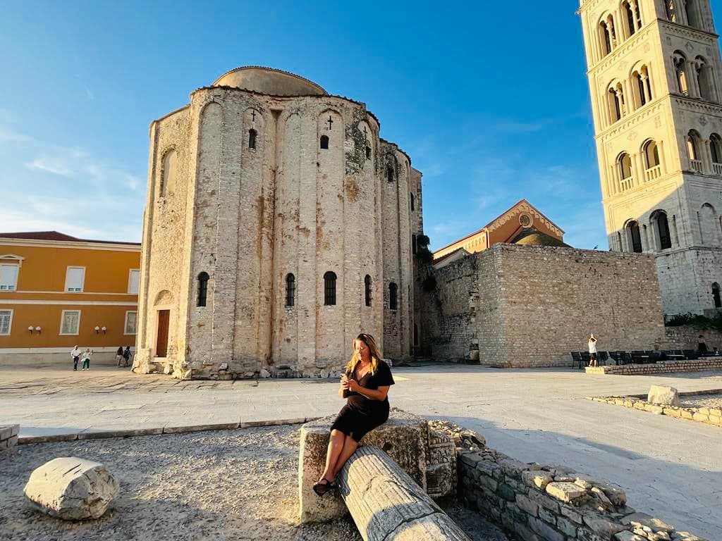 Karyn sitting in Zadar with an ancient town setting behind her and clear skies.