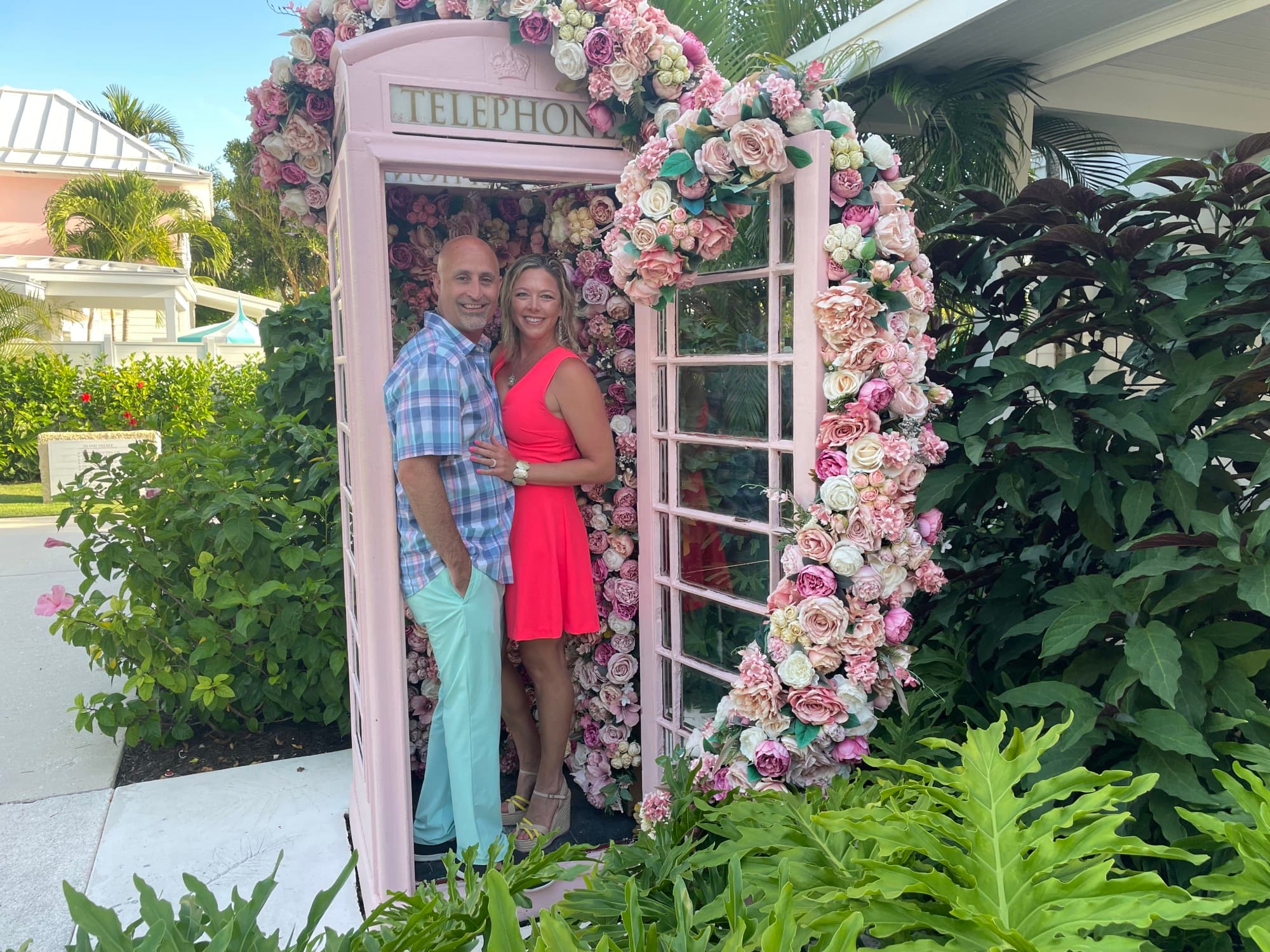 A woman in a red dress hugs a man in turquoise pants in a pink phone booth