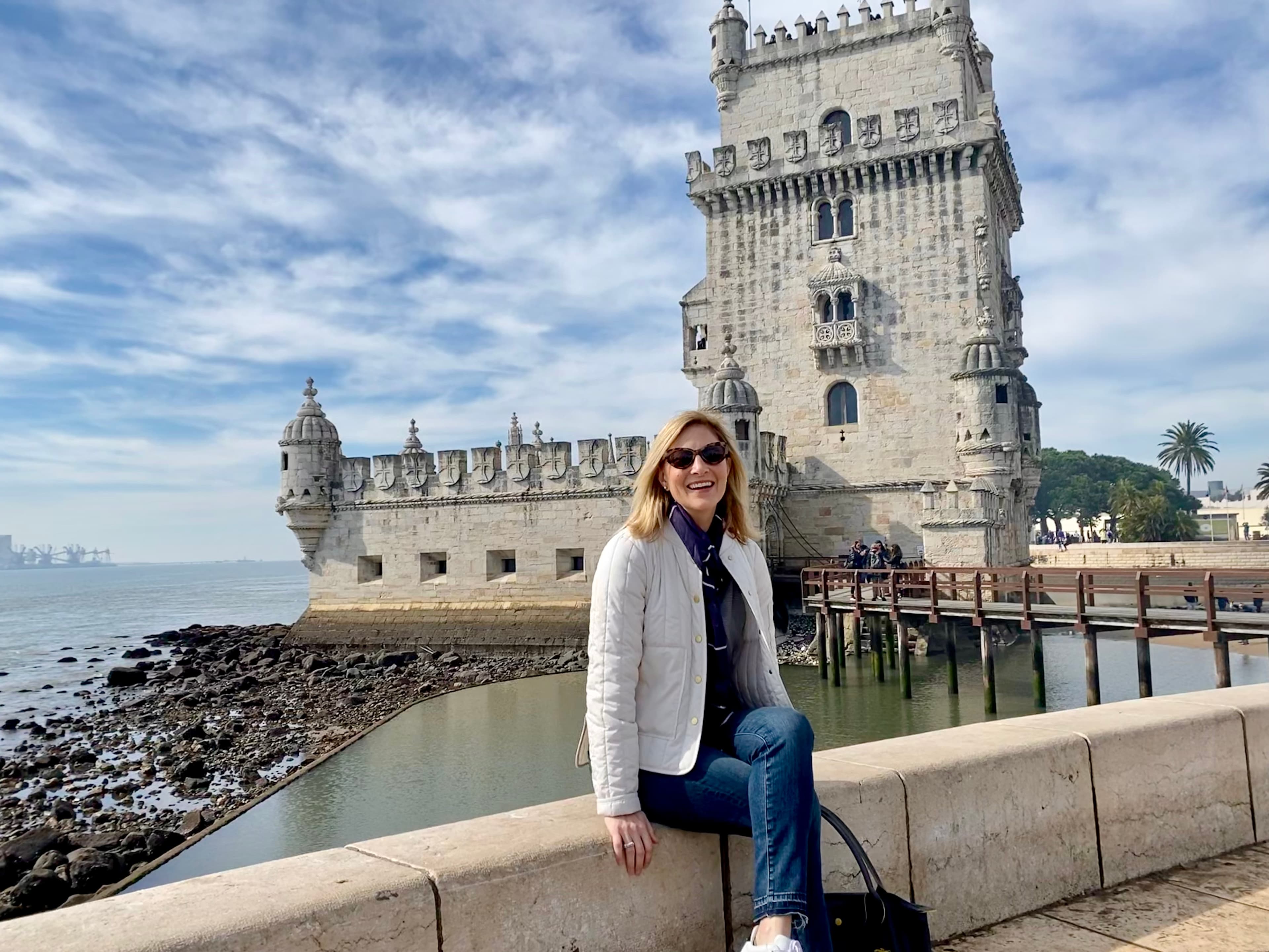 An image of Kristy wearing a white coat and sitting on a stone ledge in front of the Belém Tower