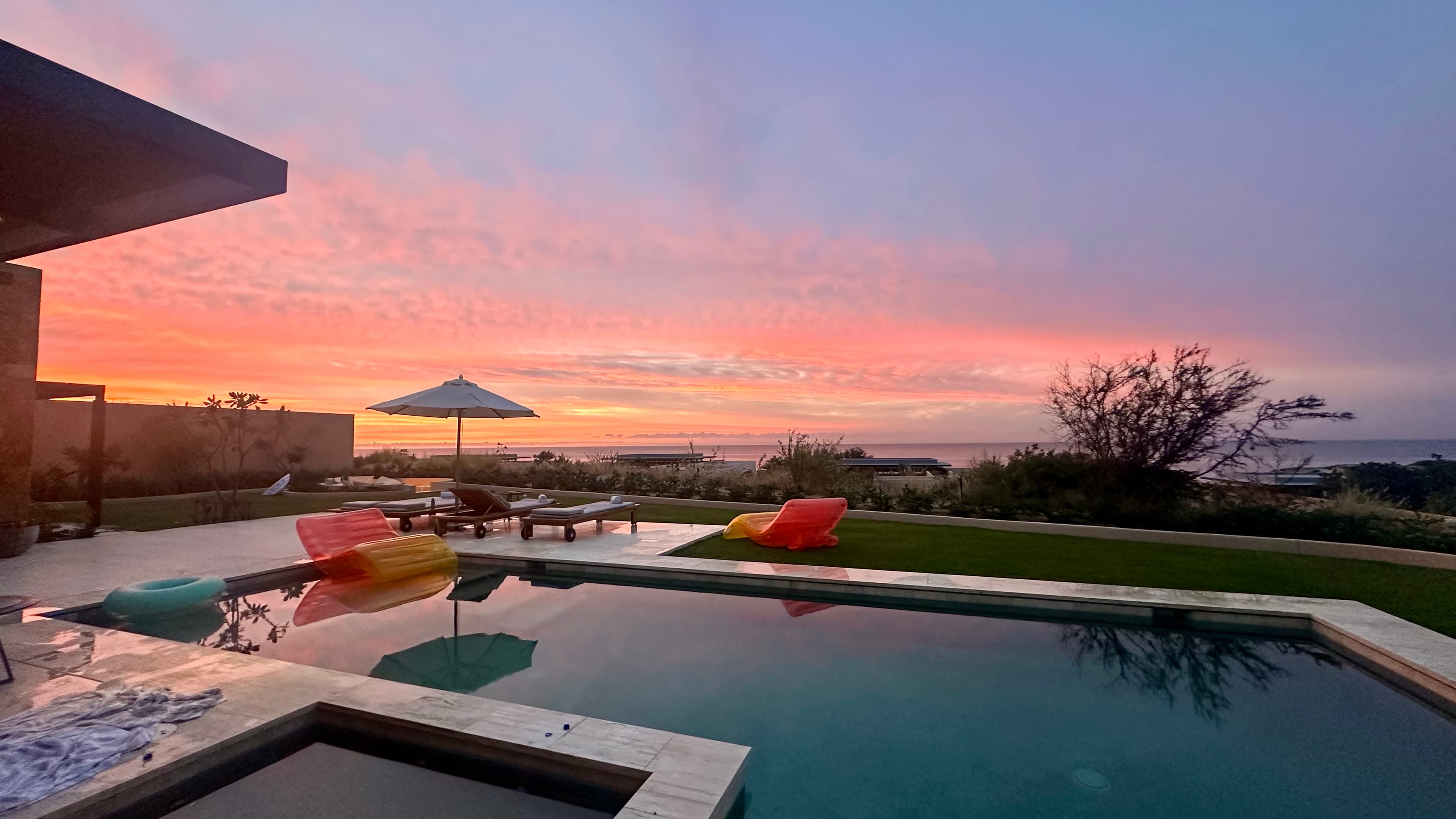 A swimming pool with inflatable lounge chairs on the water, an umbrella, lawn chair and trees with a pink and orange sunset in the distance.