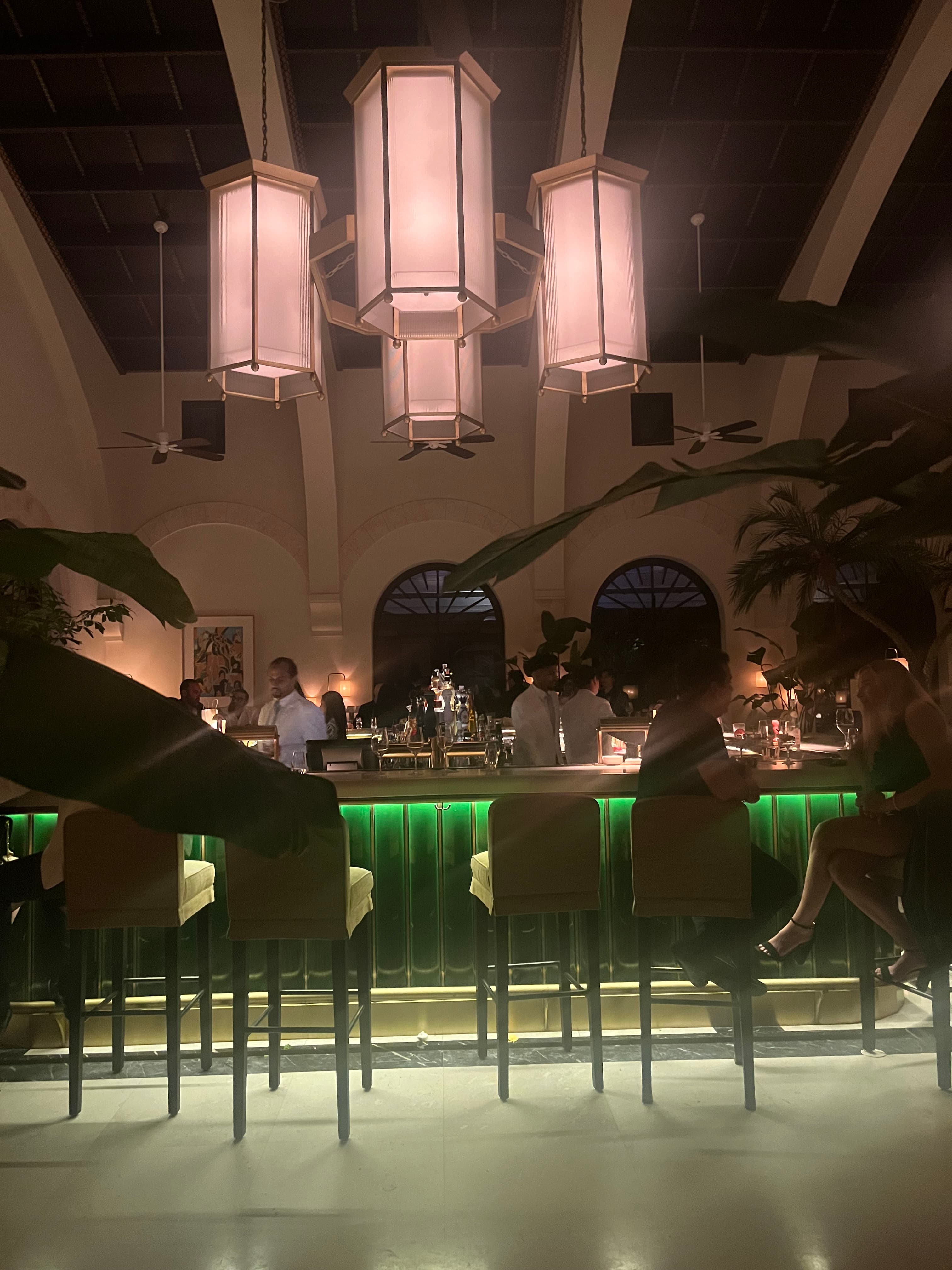A photo of a bar with green tiling, bar stools and chandeliers hanging from the ceiling