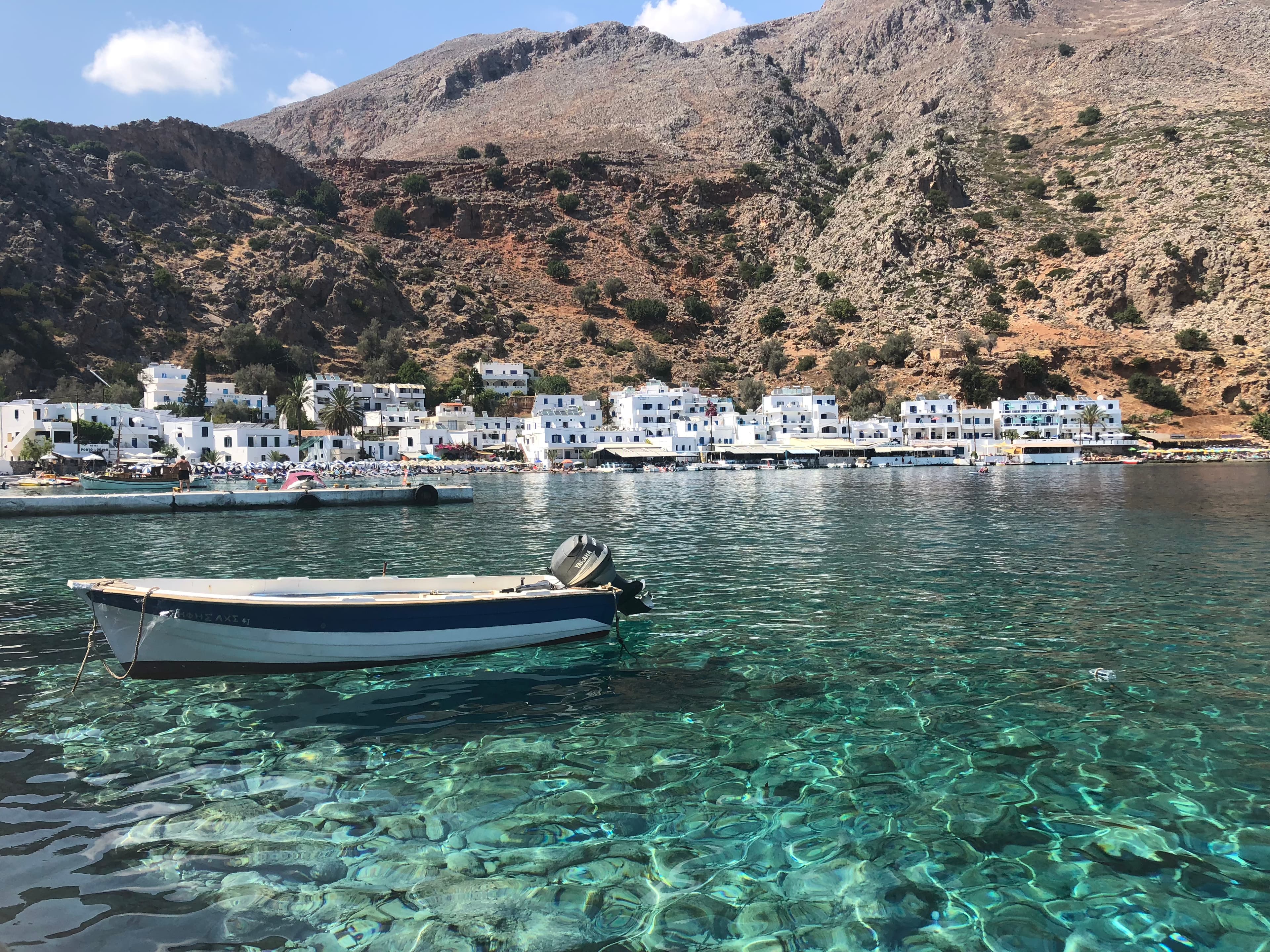 A boat floating on crystal blue water with greek buildings and a mountain in the background