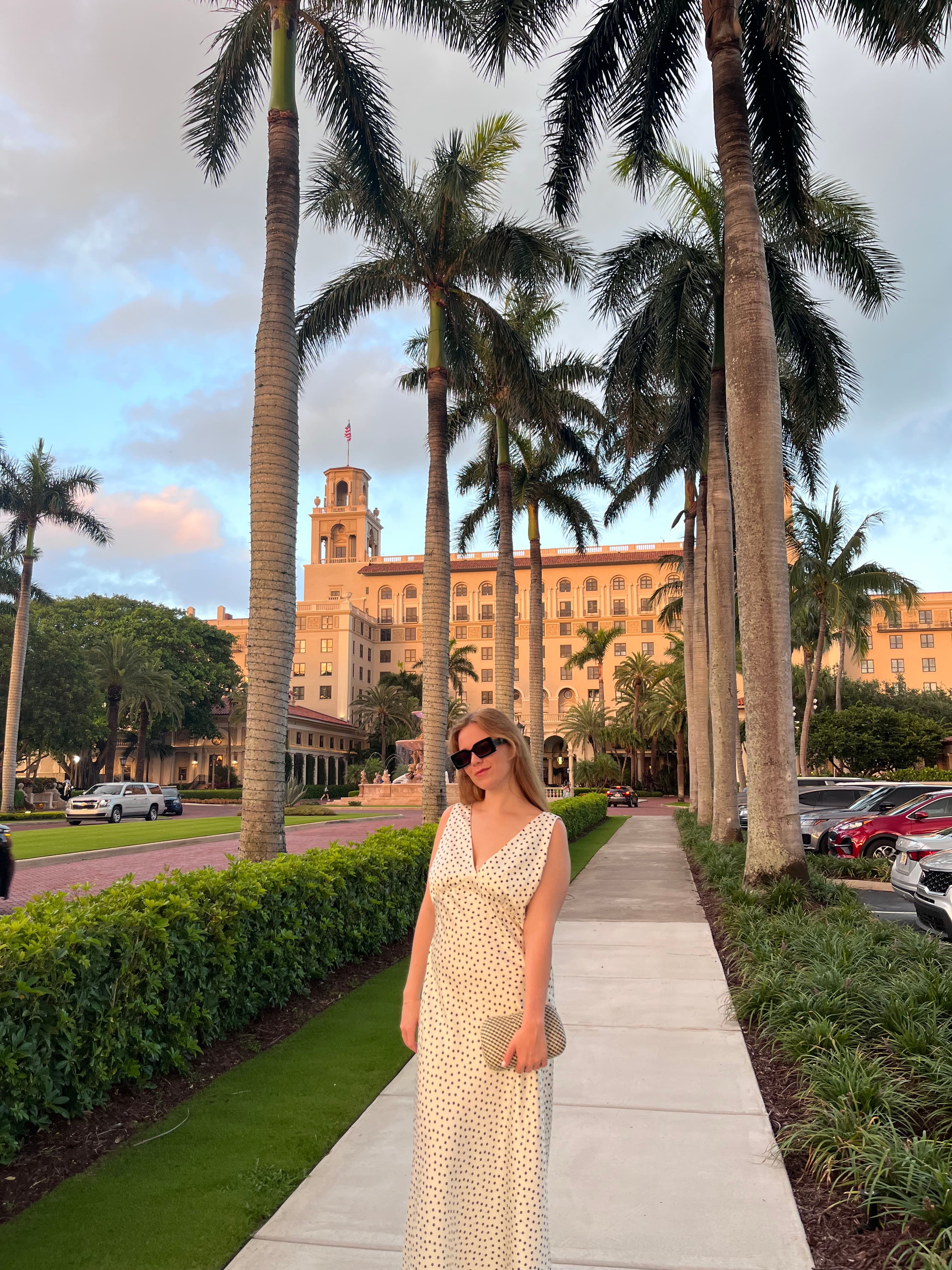 Sedona wearing a yellow dress and standing on a sidewalk in front of a hotel and palm trees
