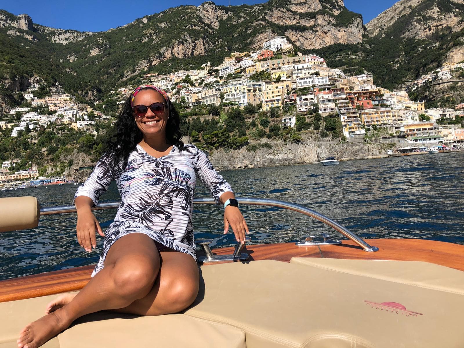 Shana sits on the front of a motor boat on the water of a coastal cliffside village