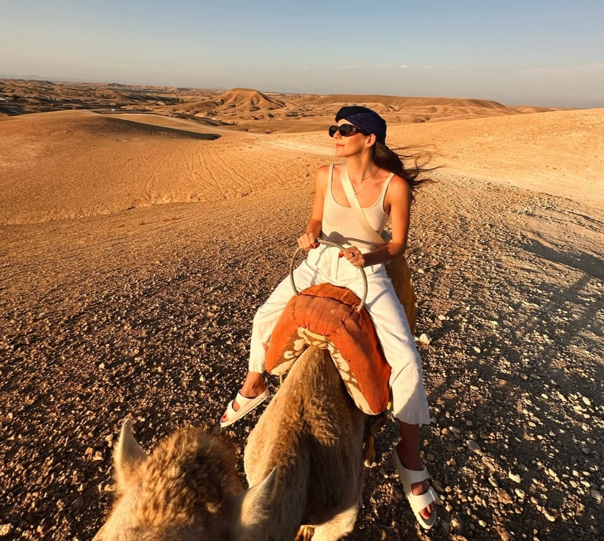 Gabby wearing a white outfit and sunglasses riding a camel in the desert