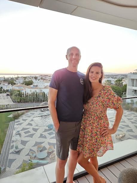 A picture of Meredith wearing a floral dress and posing with her spouse in front of a patio view at sunset