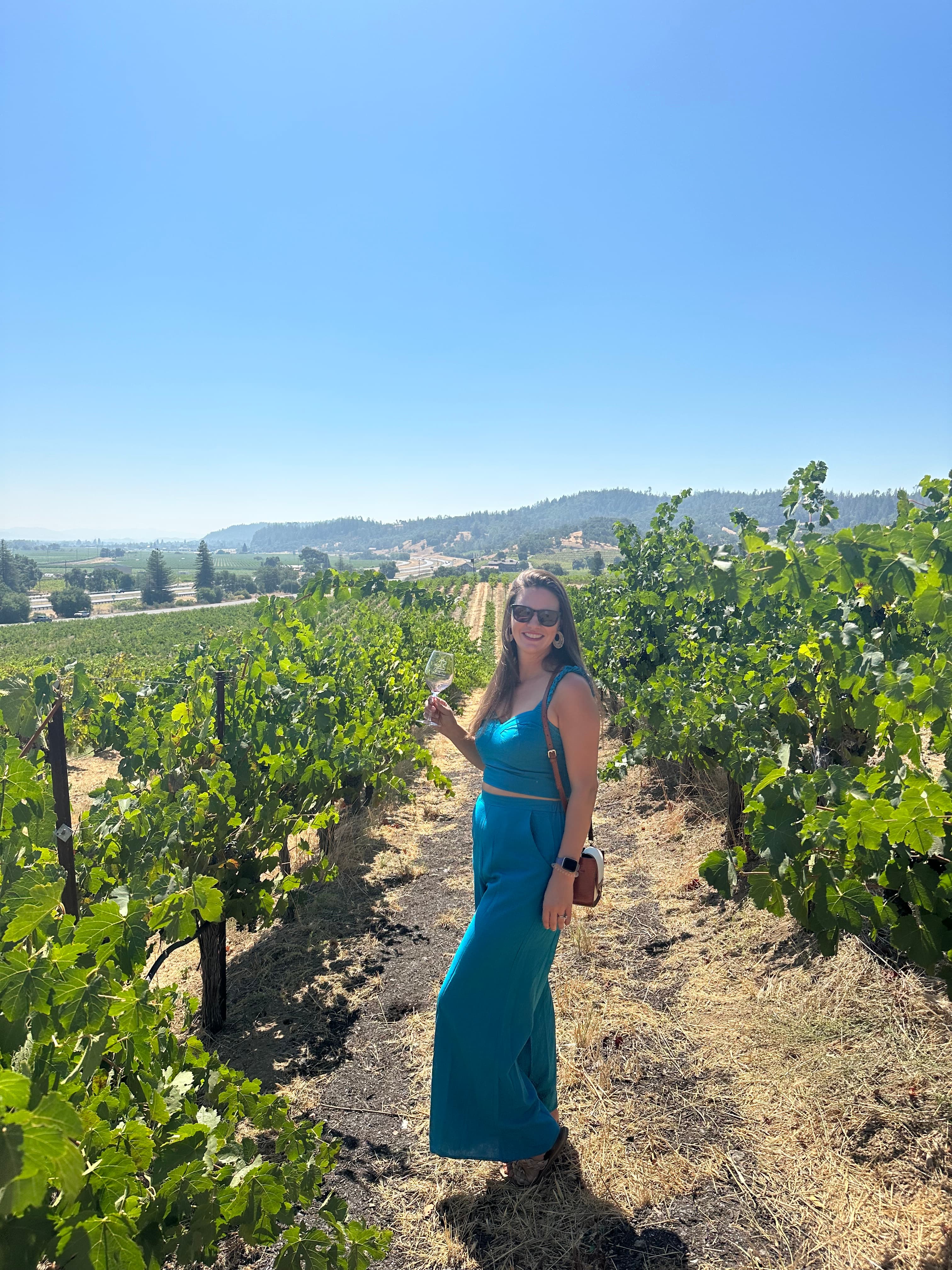 A picture of Meredith wearing a blue dress and posing on a dirt path surrounded by vineyard vines on a sunny day