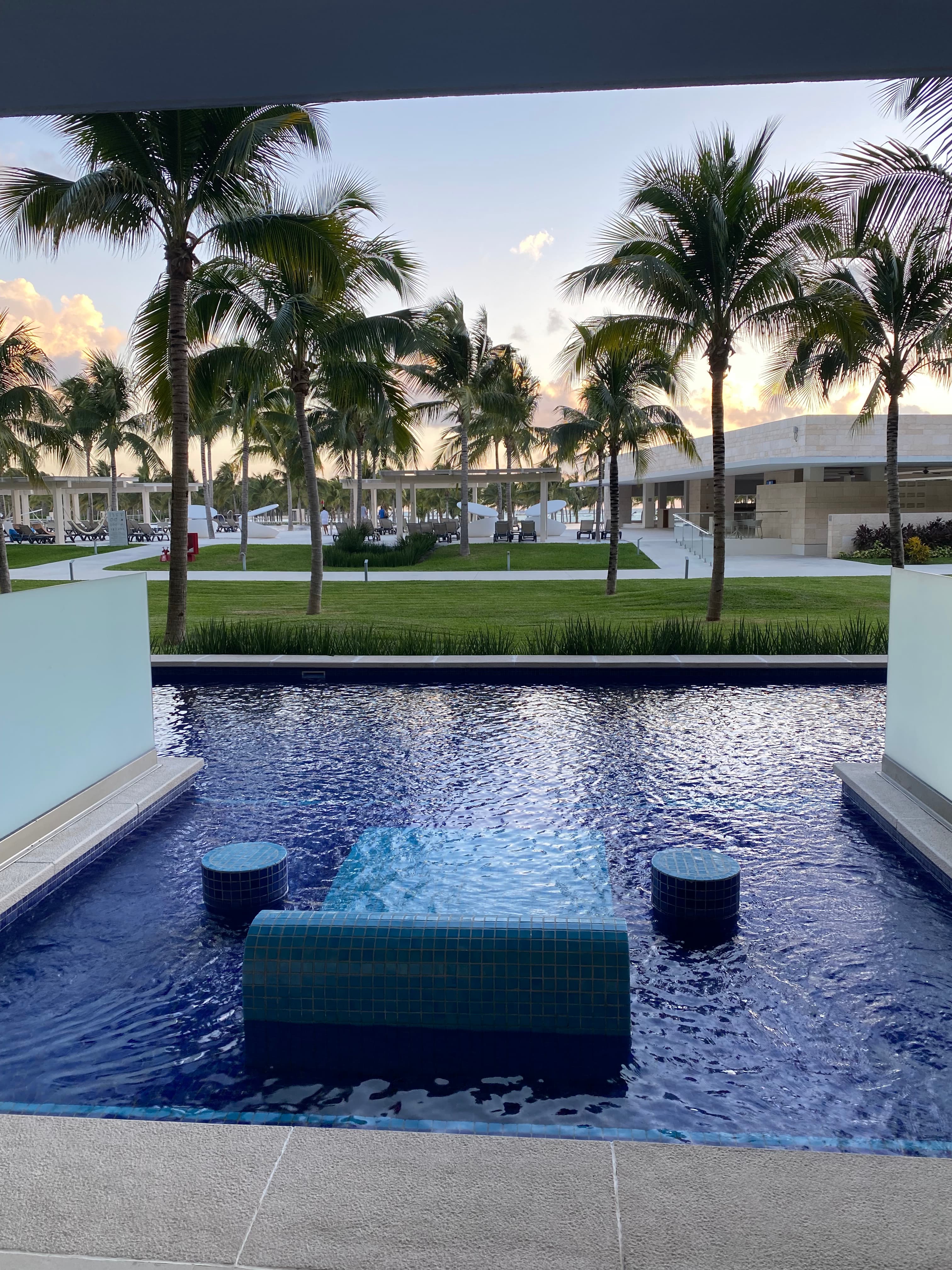 A picture of a swimming area with seating options in the water at a hotel surrounded by palm trees and green grass