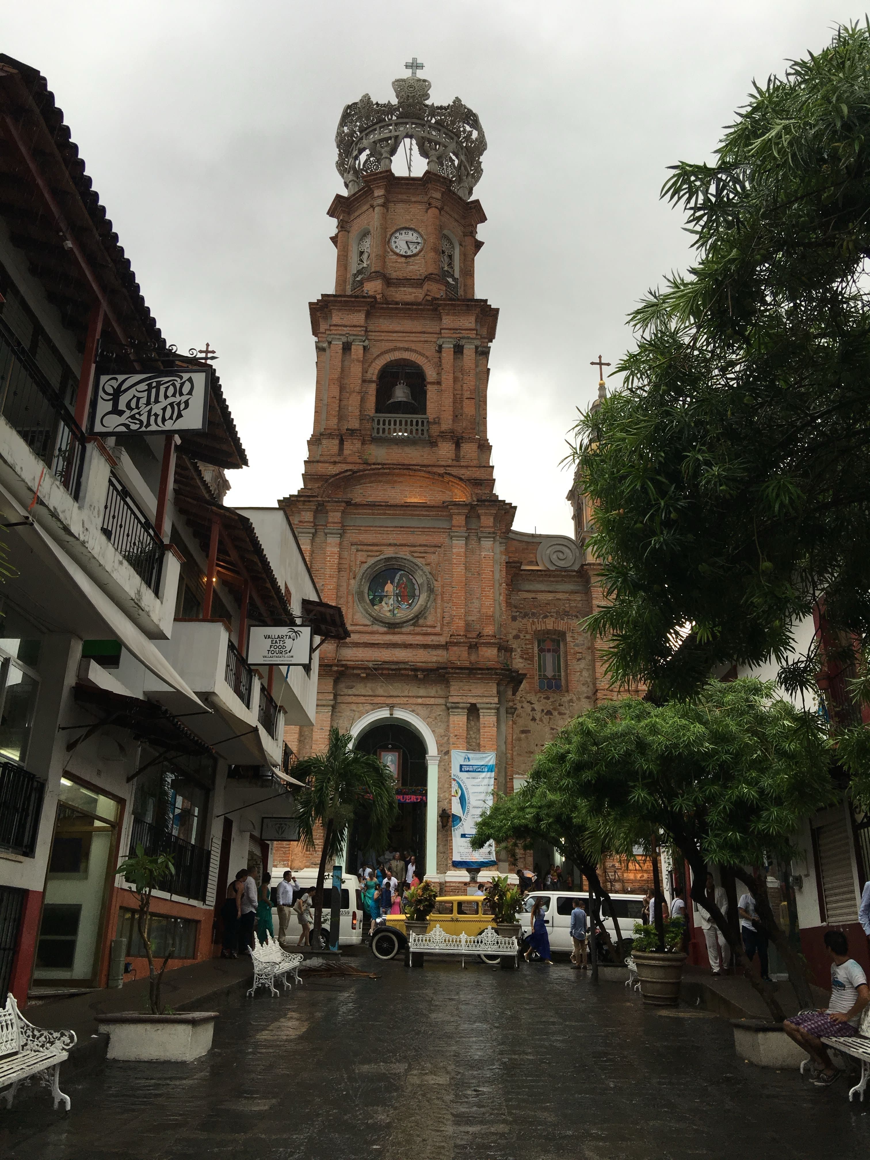A church tower in the middle of a city square surrounded by buildings and green trees on a cloudy day
