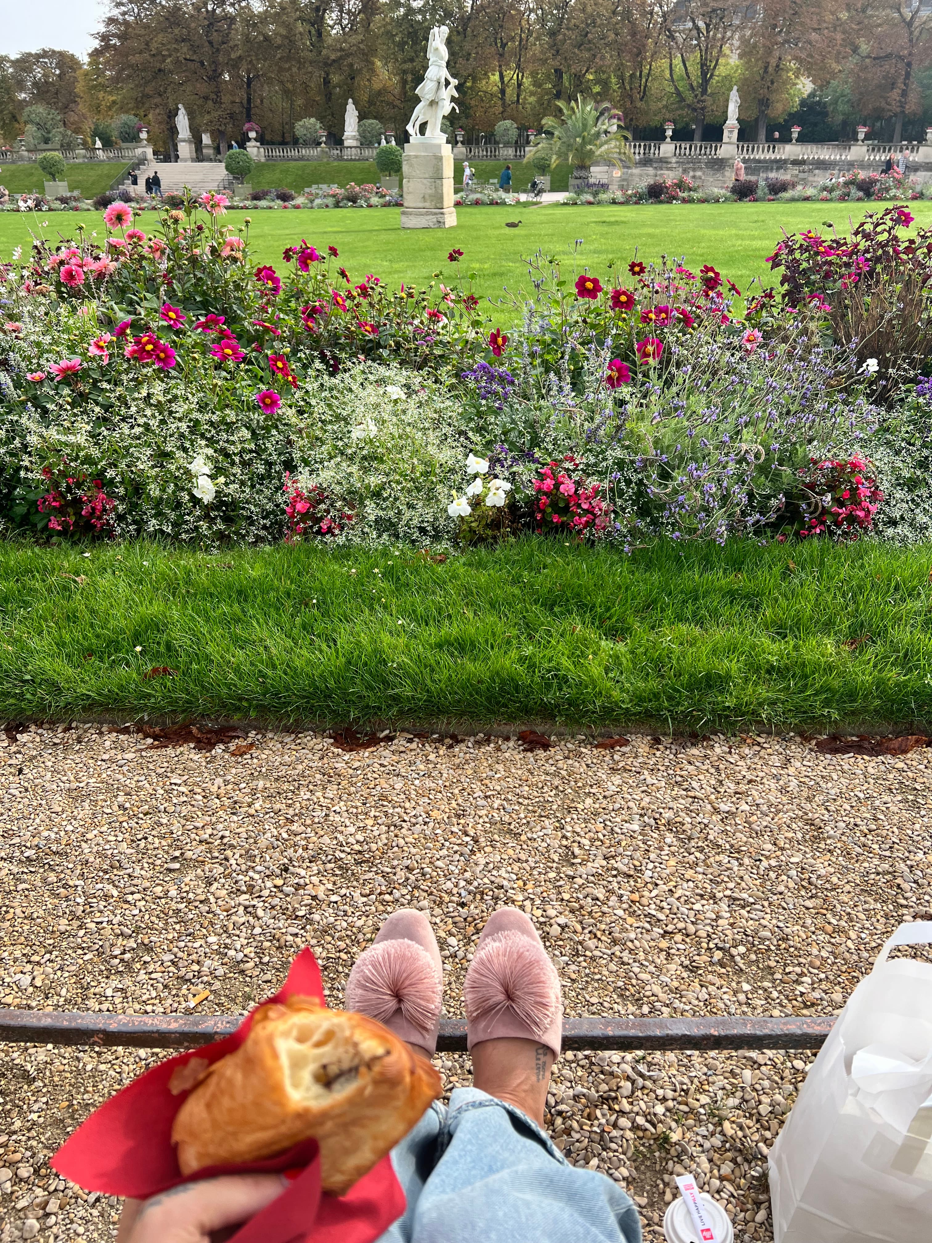 Amanda seated while holding a pastry in front of a dirt path and garden with statues in the distance