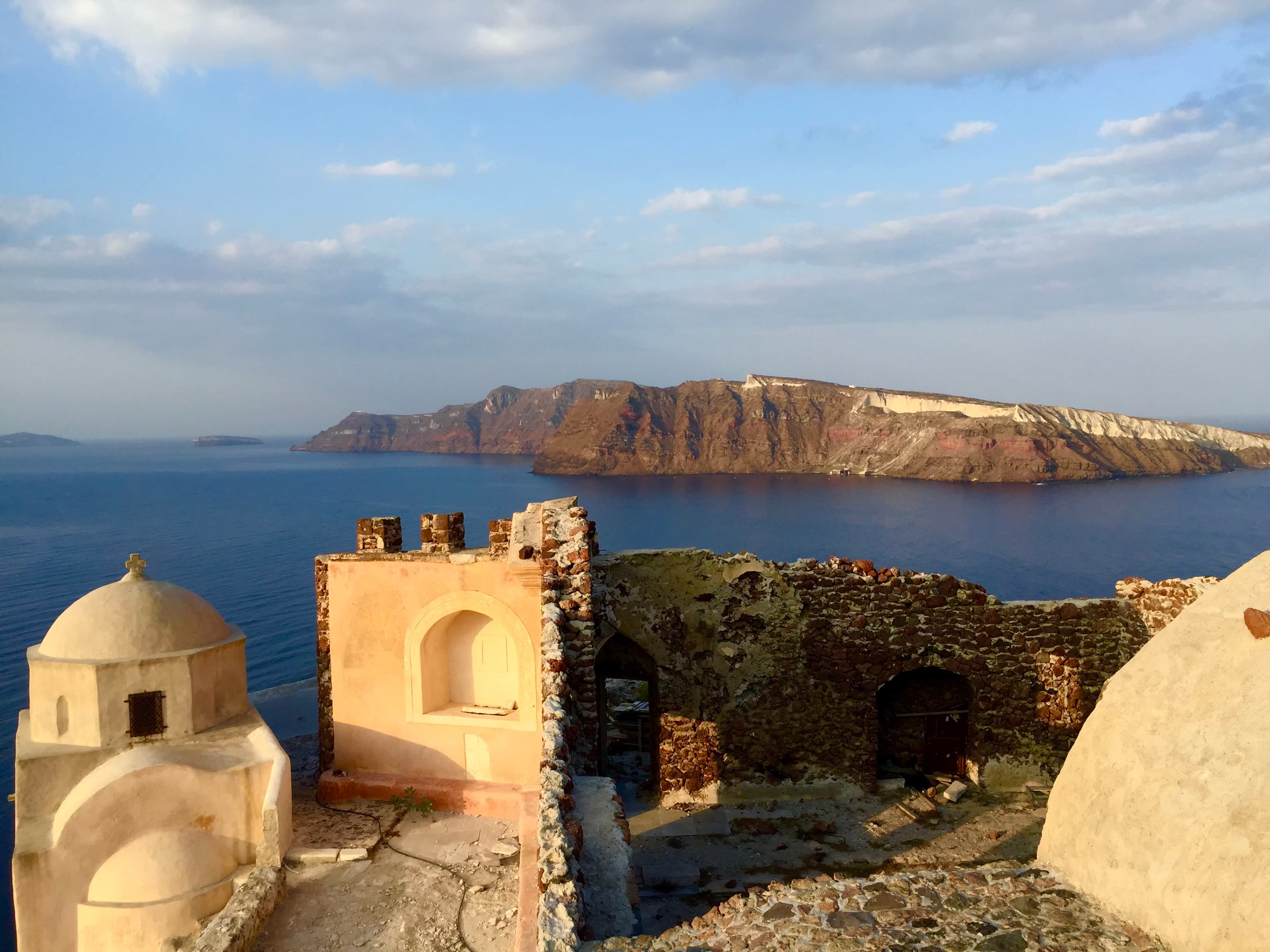 A sunlit view of Greek buildings with blue water and mountains in the background