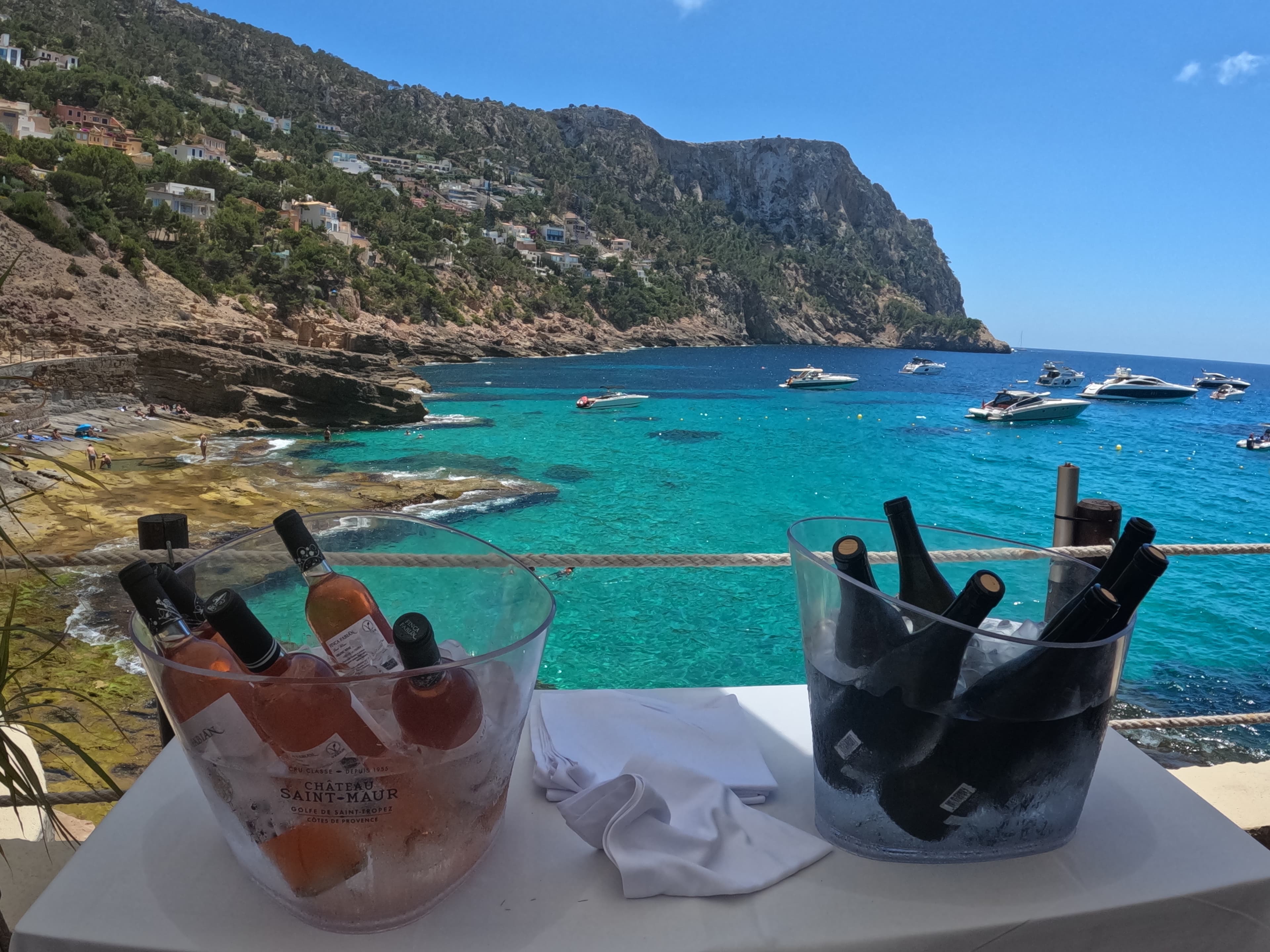 Two buckets of drinks on a white table in front of a turquoise blue body of water and green mountain.