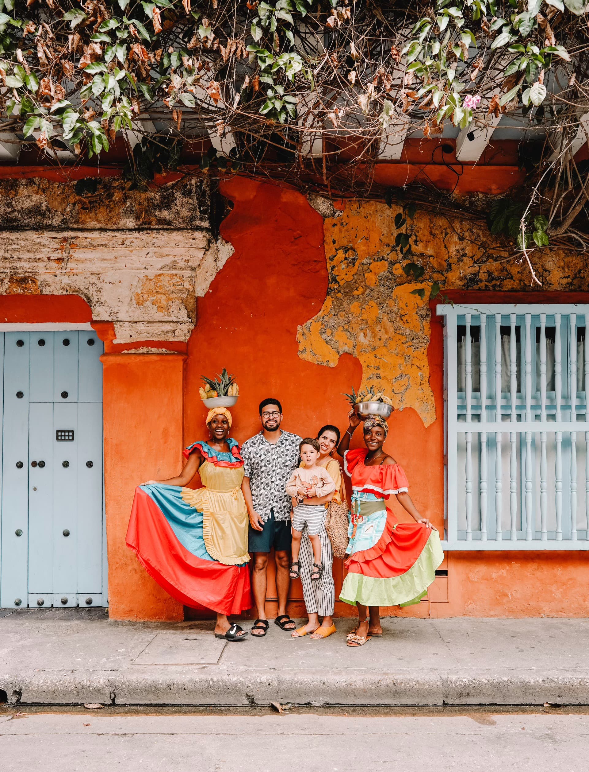 Maria, husband and baby posing for a photo with locals wearing traditional clothing in front of a bright and colorful building.