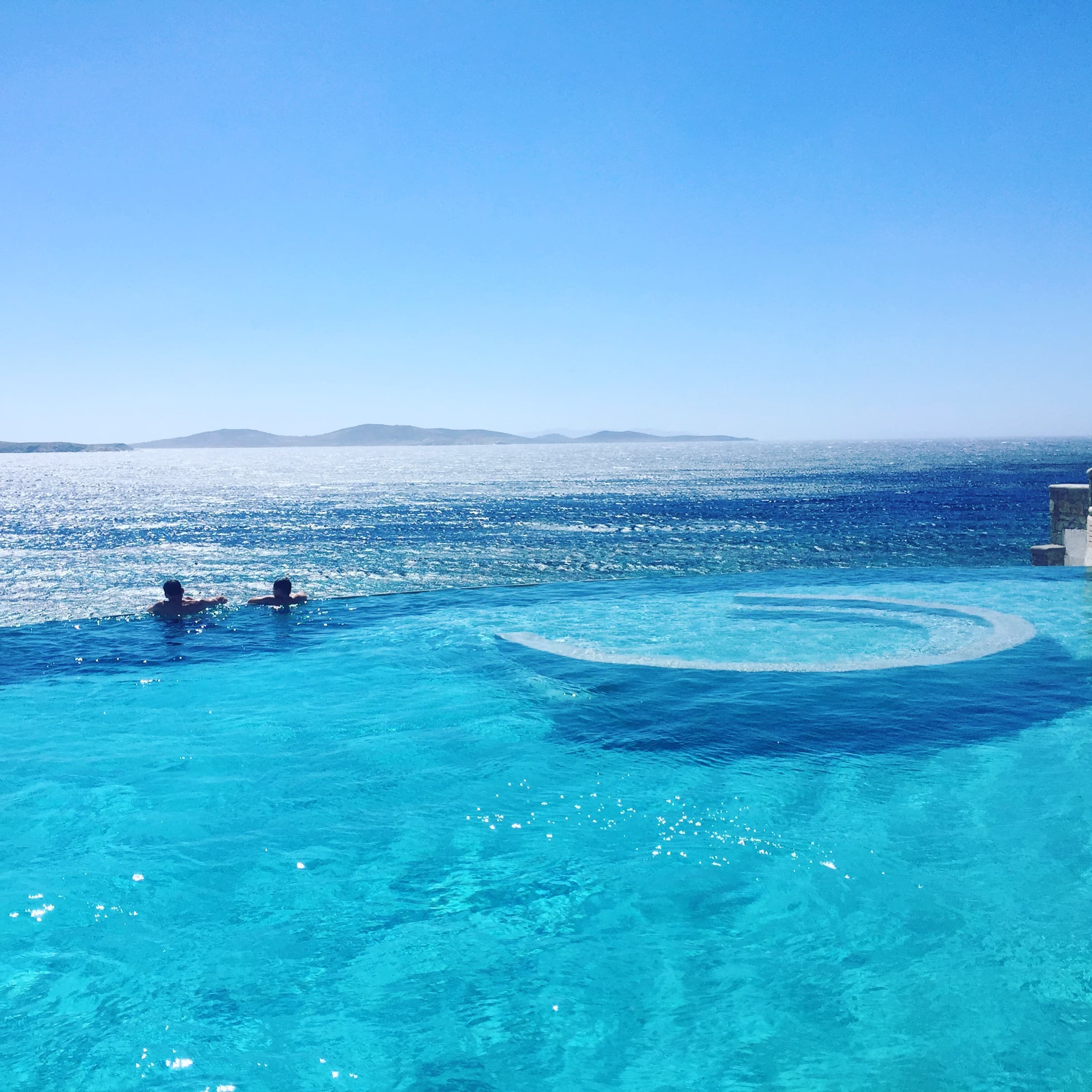 A photo of a blue pool with two people in the water overlooking the sea with the sun sparkling overhead
