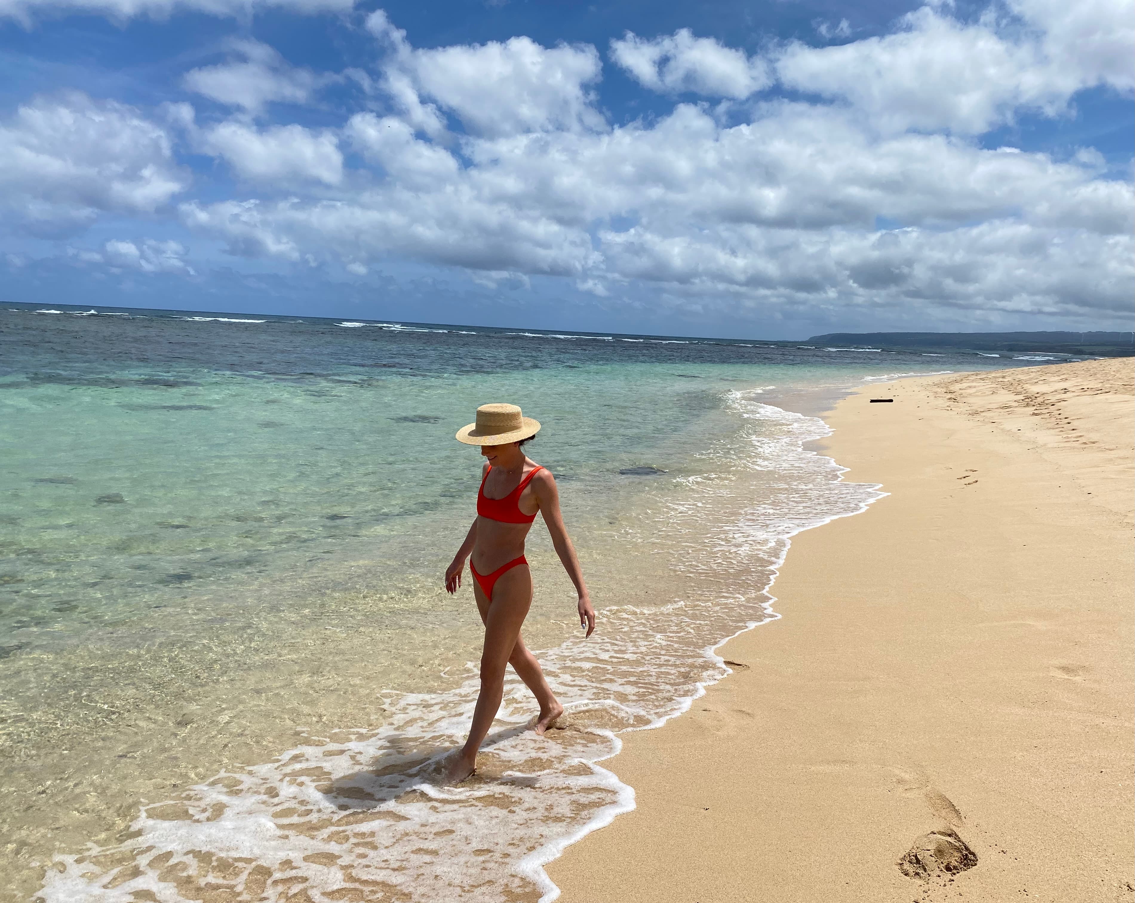 Gabby wearing a swimsuit and hat walking on the beach with the ocean and cloudy blue sky in the background