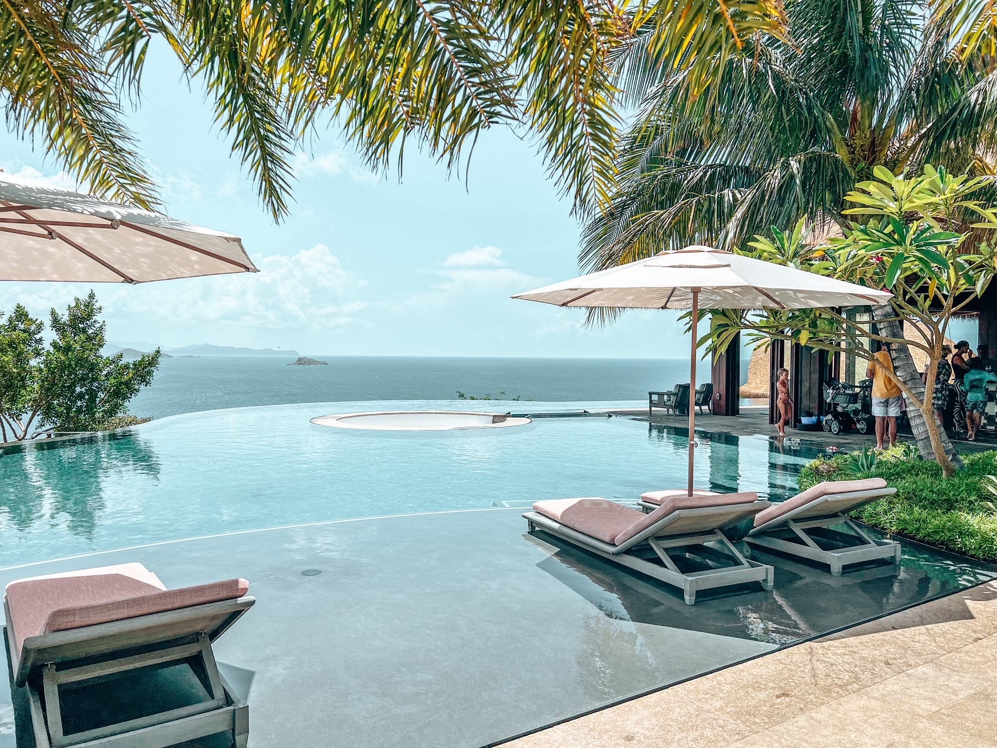 Three lawn chairs and two umbrellas positioned poolside and surrounded by palm trees looking out to the sea