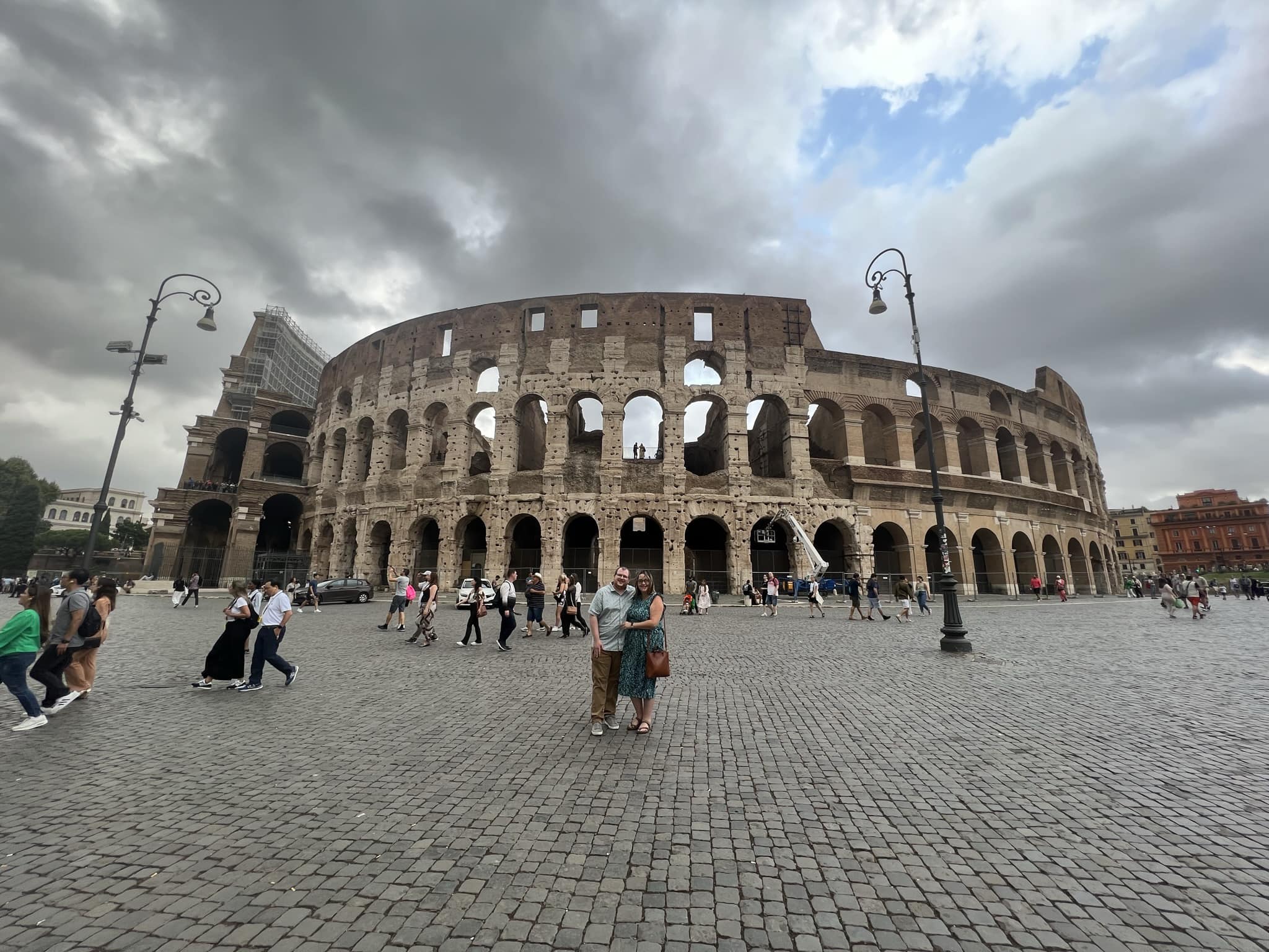 Couple posing in front of the Colosseum in a large open pedestrain area with many people in Rome
