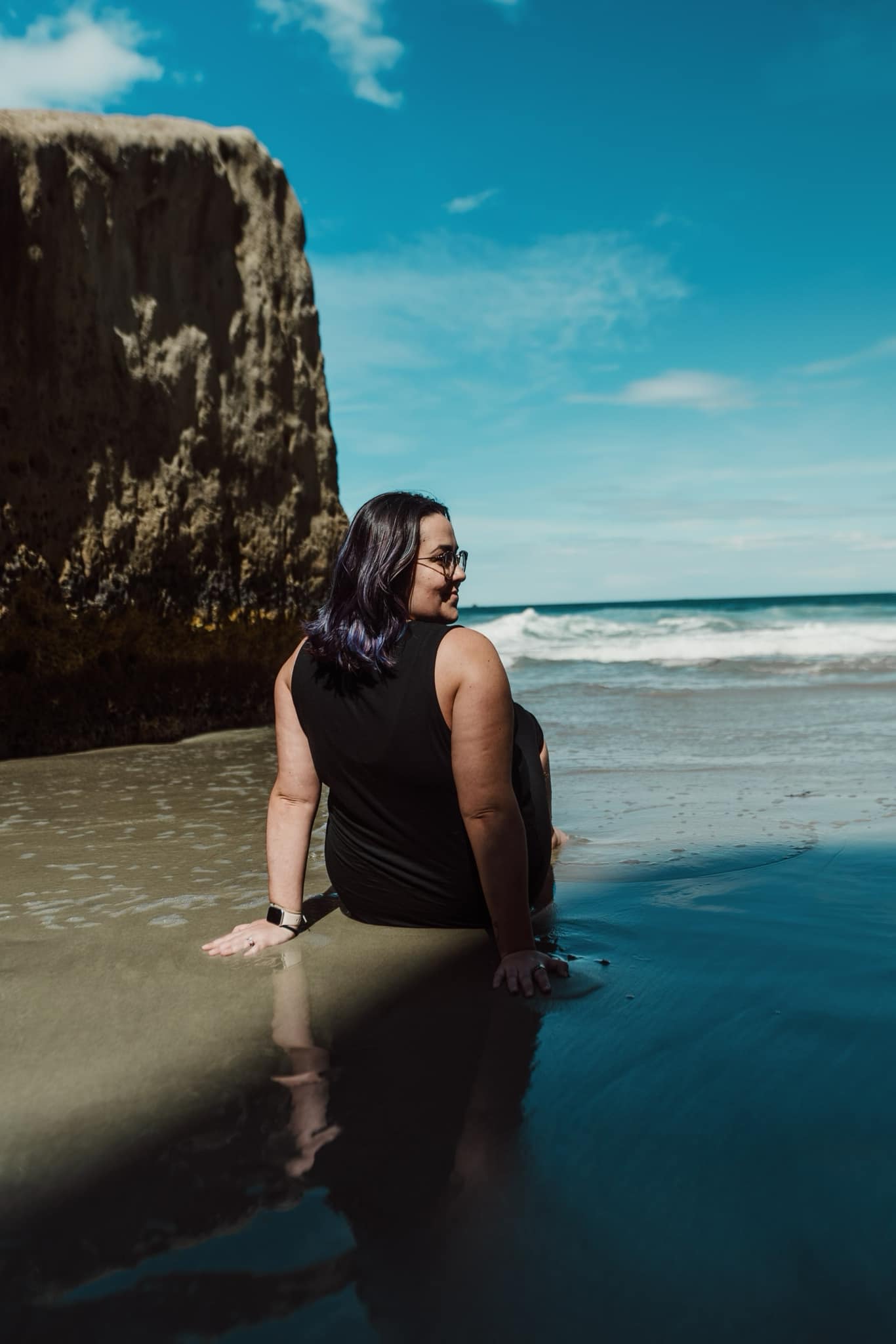 Rear view of women sitting on the sand on a beach in a black top with a large rock formation in view
