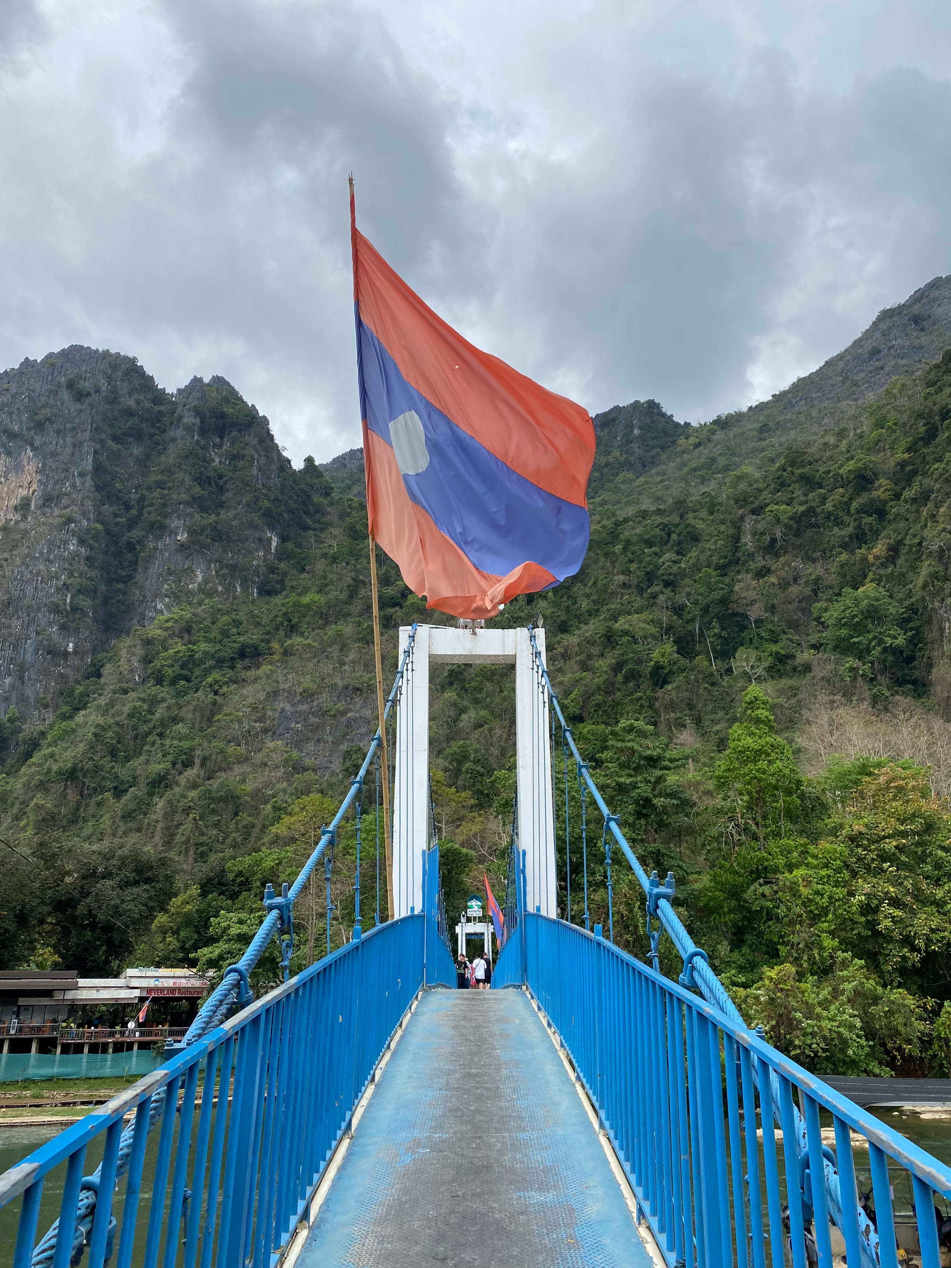 A photo of a blue bridge with mountains in the background and the Laos flag flying above it.