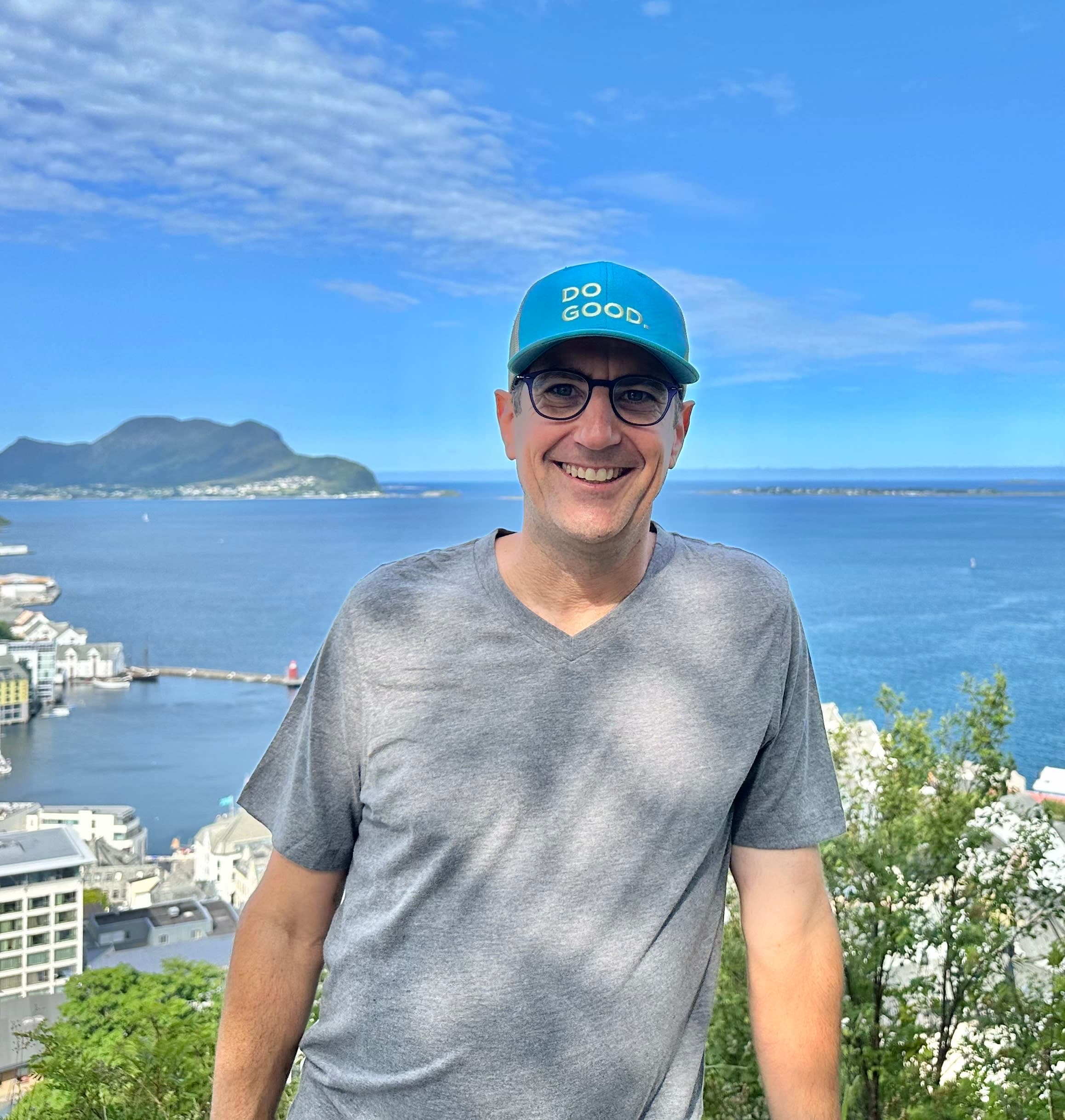 Photo of Dave in a blue cap and gray shirt posing next to a bush overlooking the sea