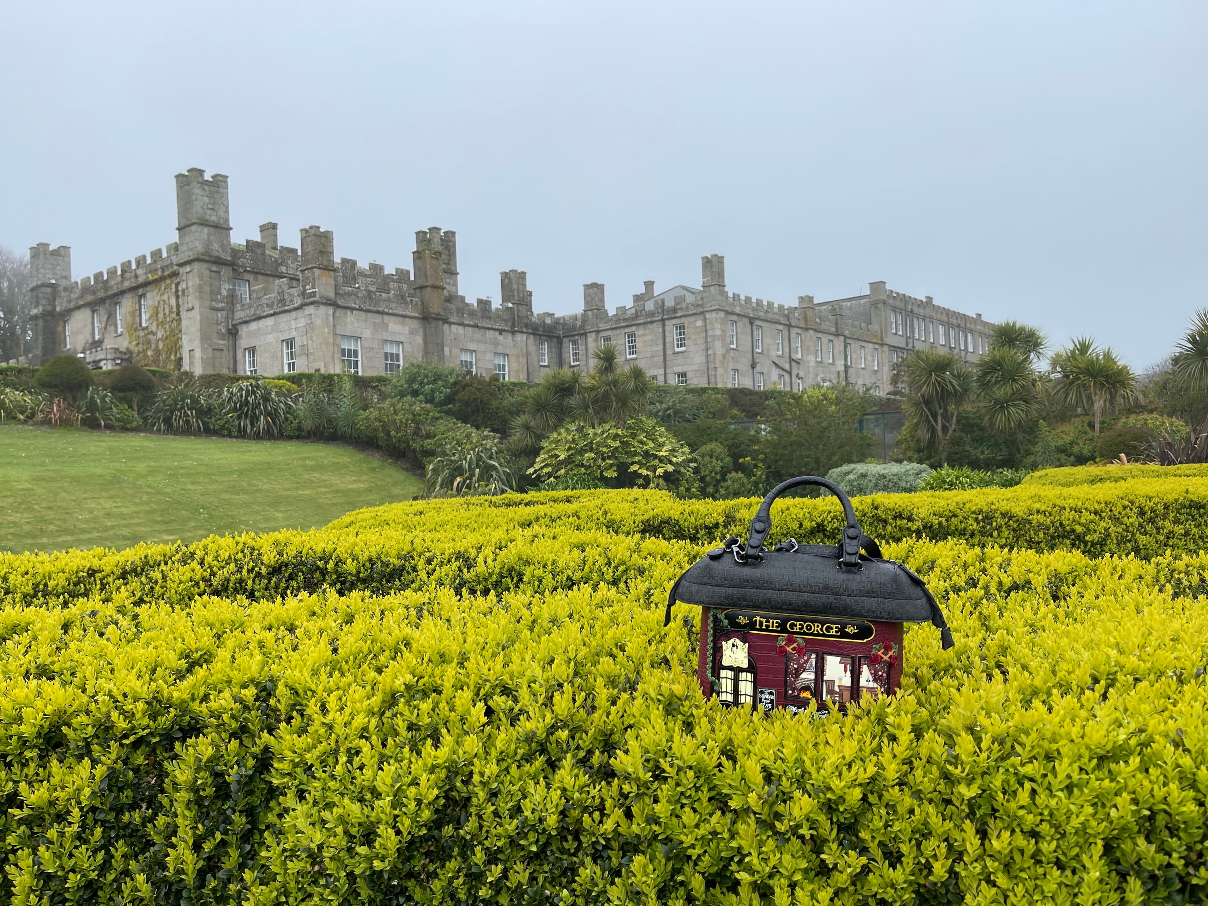 A purse decorated to look like the George pub sitting in bushes with a view of the Tregenna Castle Resort in the background.