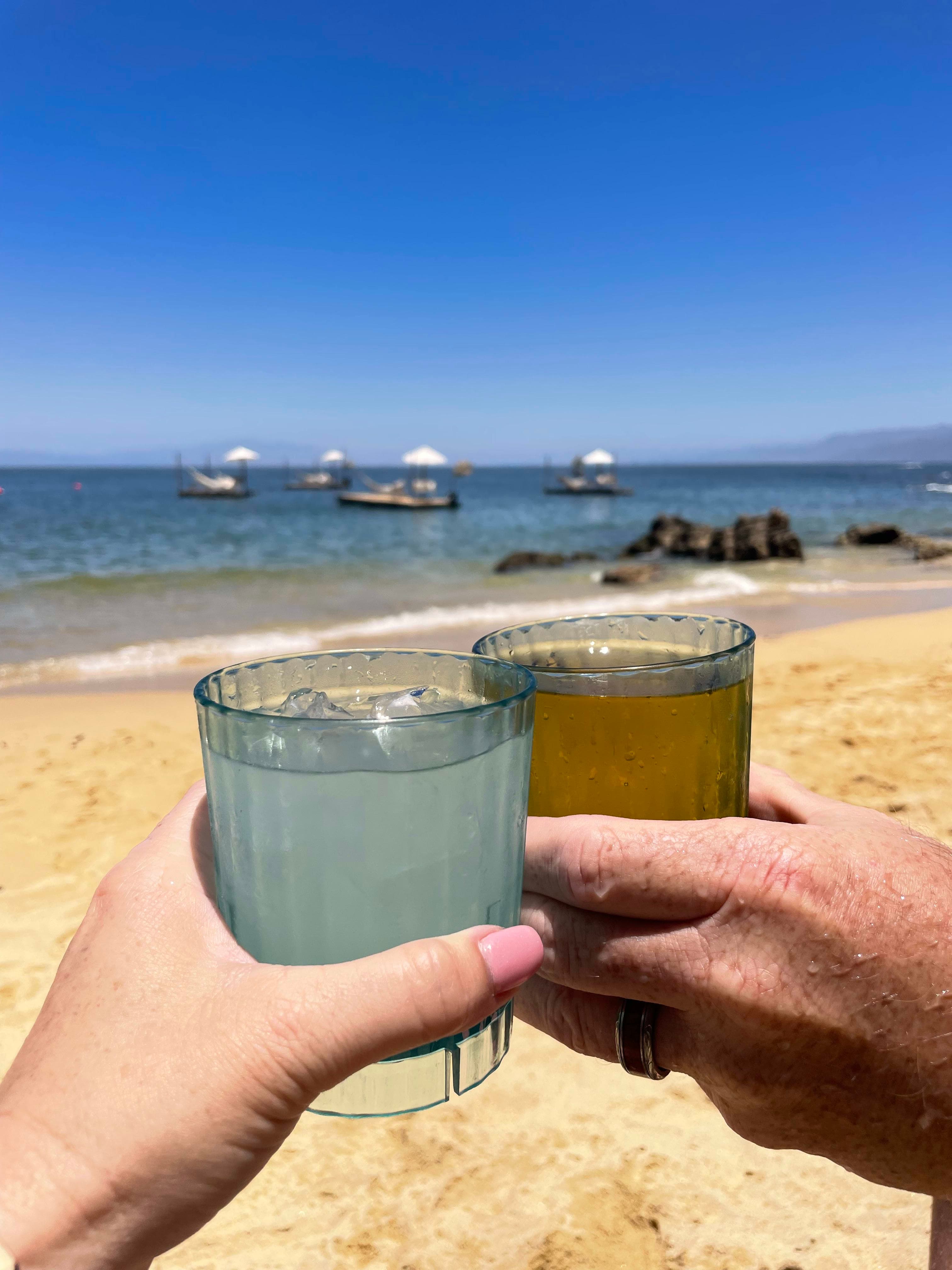 A photo of two drinks beind held on a beach