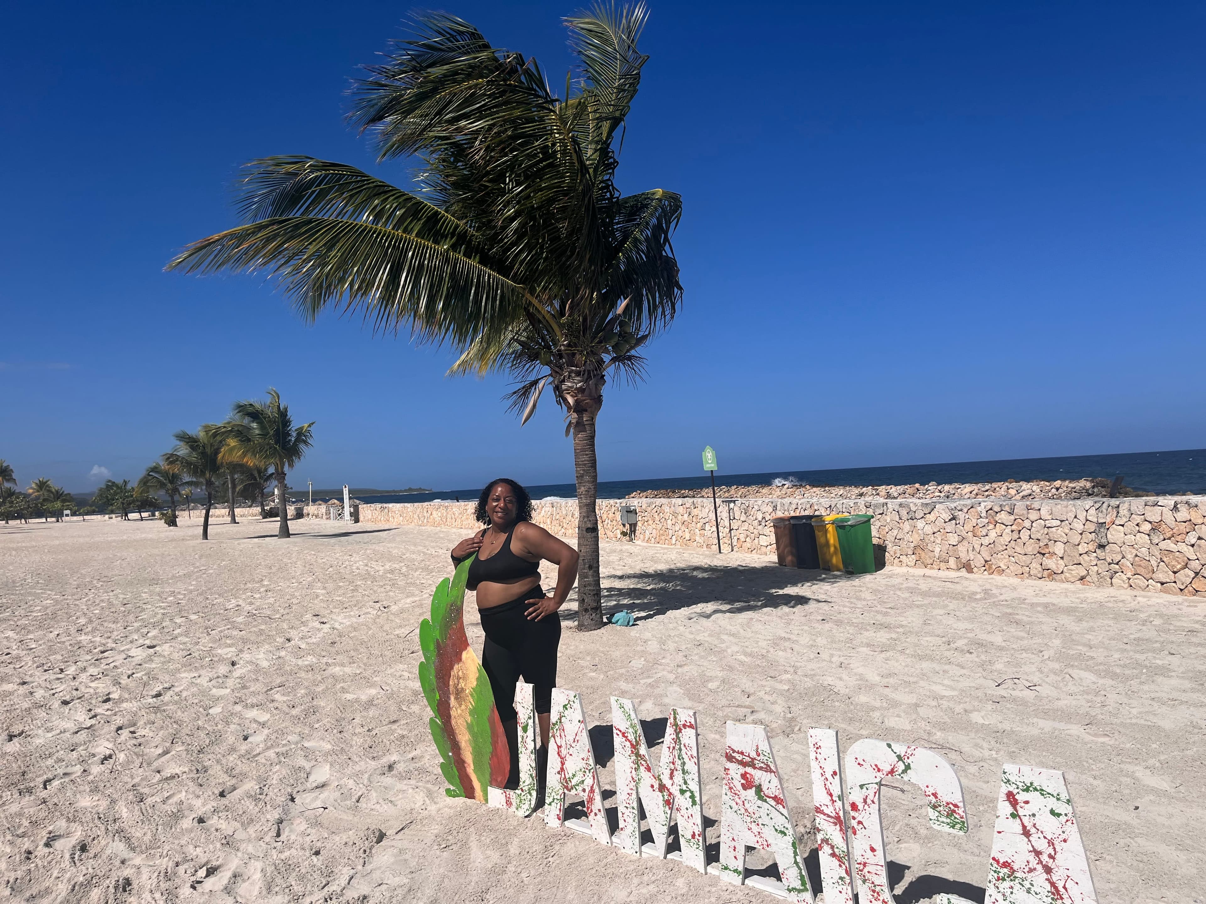Ashley posting against a sign that says “Jamaica” on a white-sand beach under blue skies