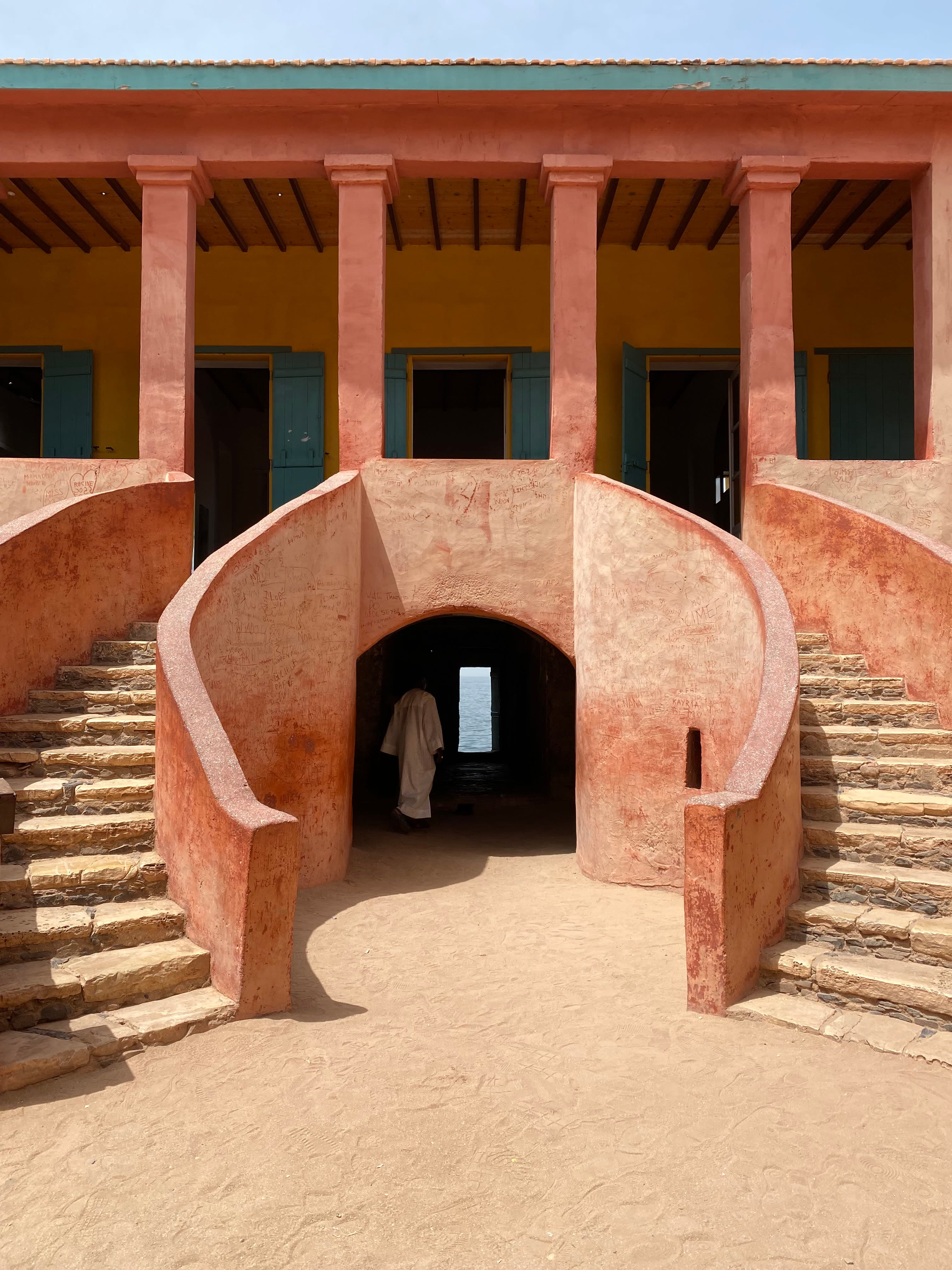 A figure in a white gown, standing beneath an entryway of two parallel staircases, leading up to a red clay building.