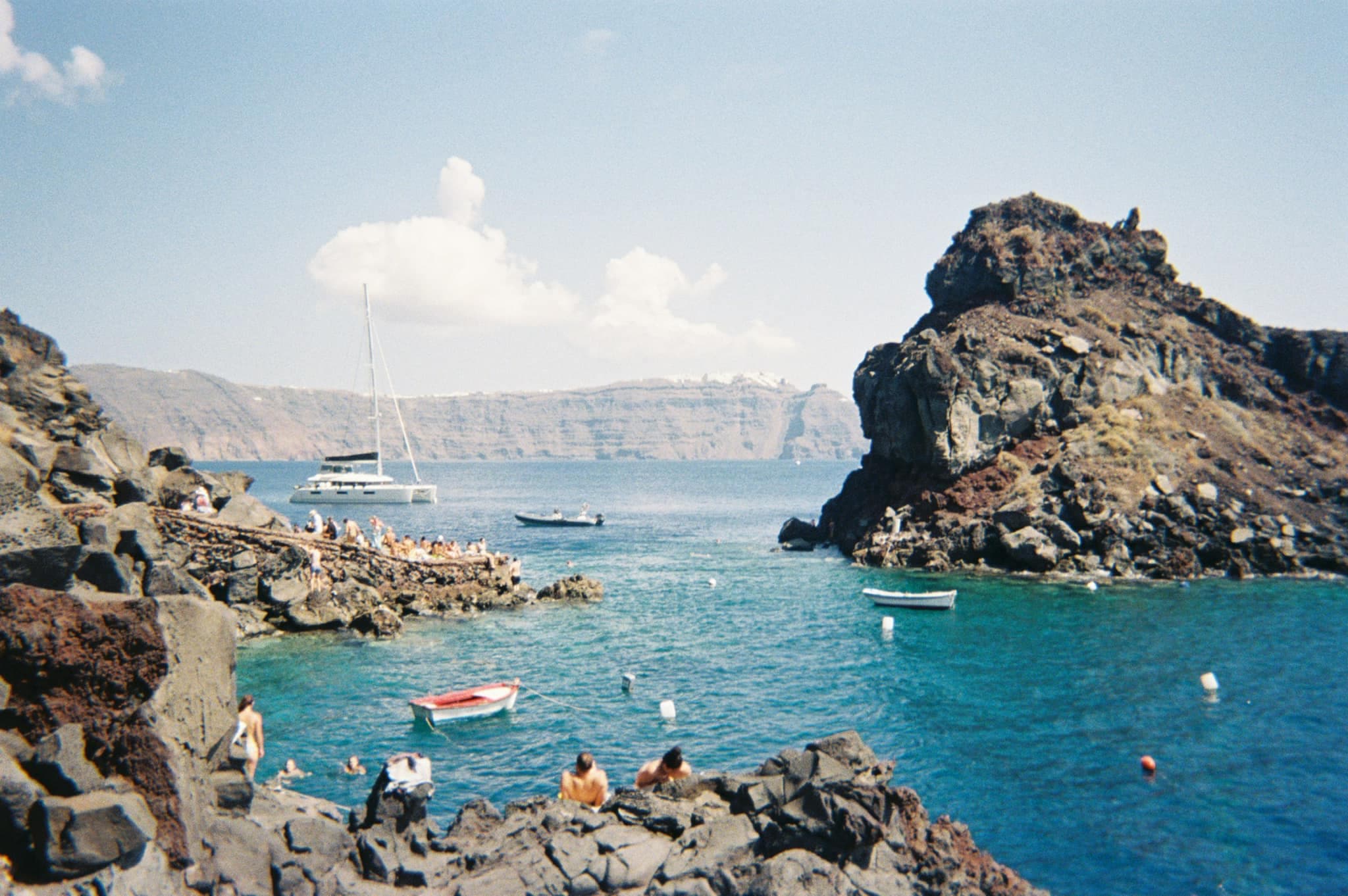 A film photo of Amoudi Santorini, a cove with rocky shores, people swimming and boats anchored in it.