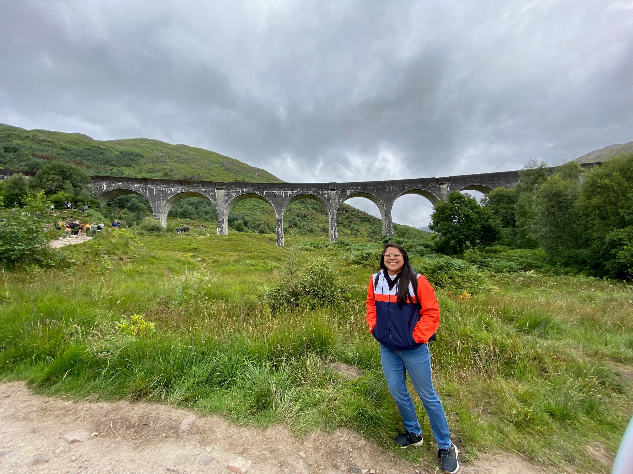 Travel advisor Alyssa standing in front of a green field with an old stone bridge with arches on a cloudy day.