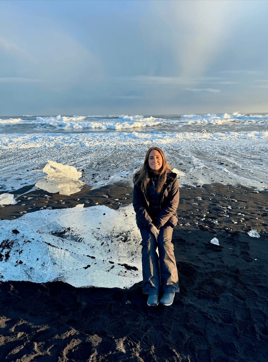 Sofia in a winter coat smiling in front of a snowy and rocky landscape under cloudy skies